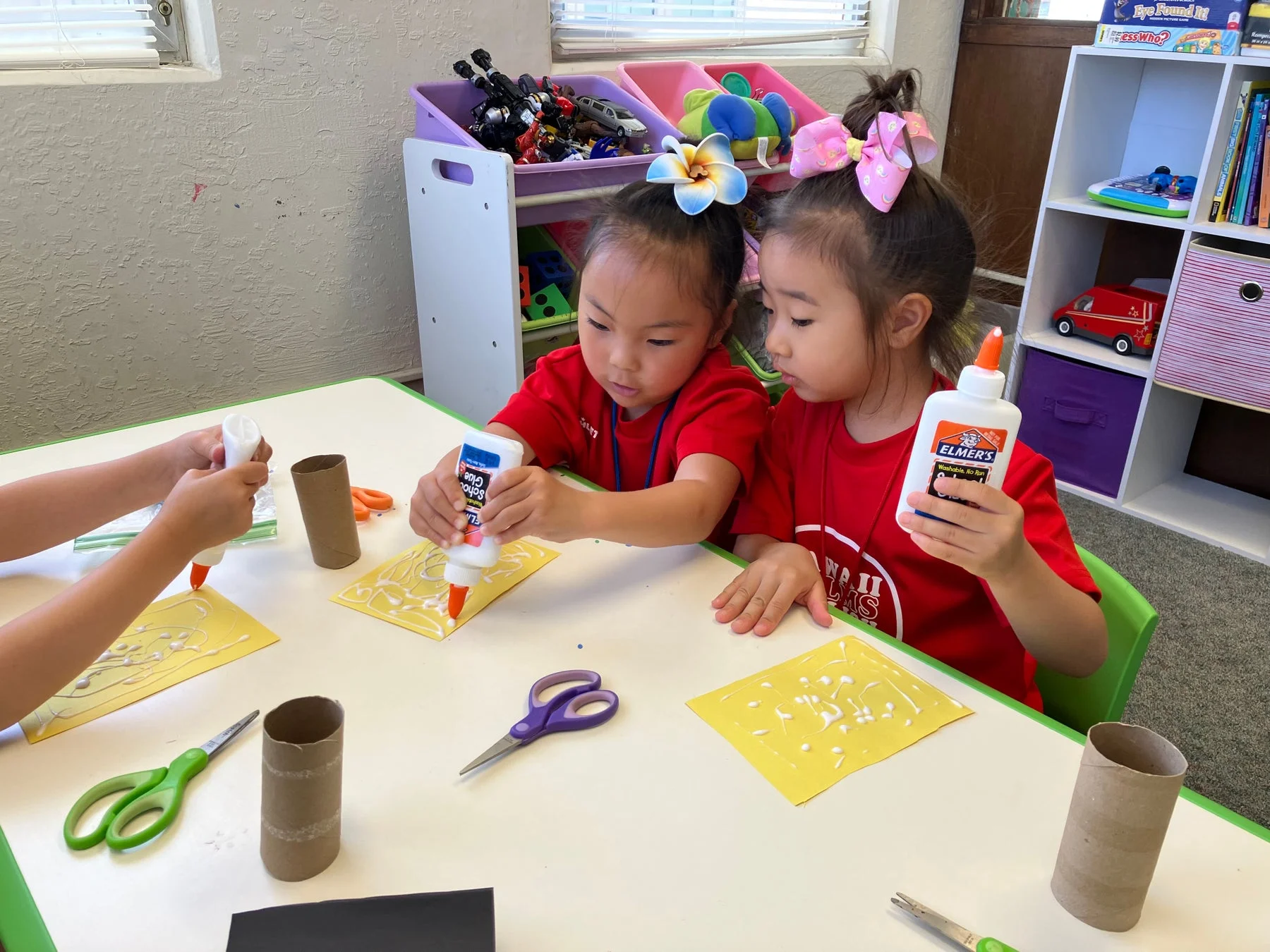 二人の幼い女の子がテーブルで工作活動に集中しています。彼女たちは赤いTシャツを着ており、のりを使って紙に模様を描いています。教室のような環境で、創造的な遊びを楽しんでいる様子がうかがえます。
