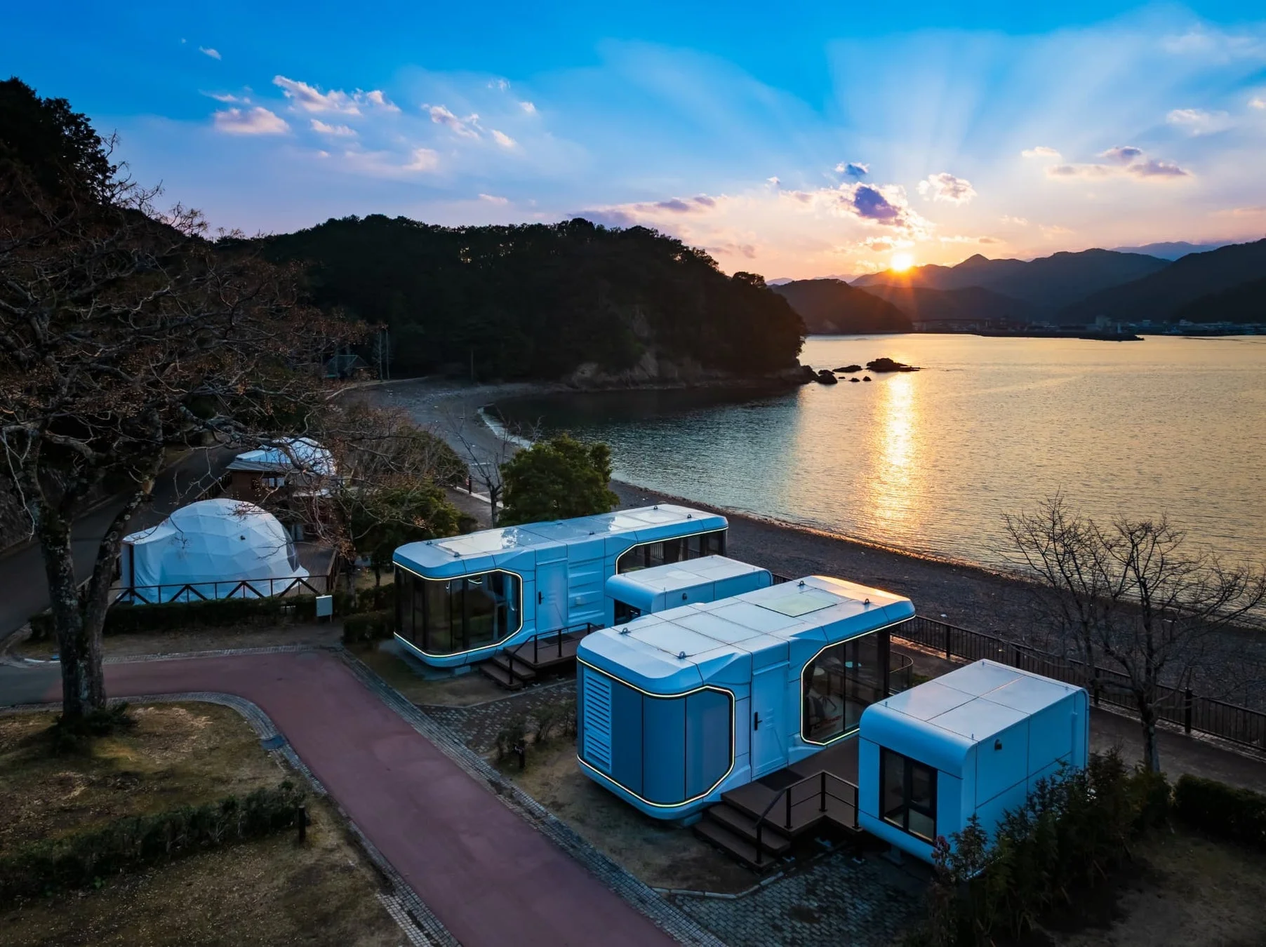 夕暮れ時、海辺に並ぶモダンなモジュール型宿泊施設とドーム型テントのグランピング風景