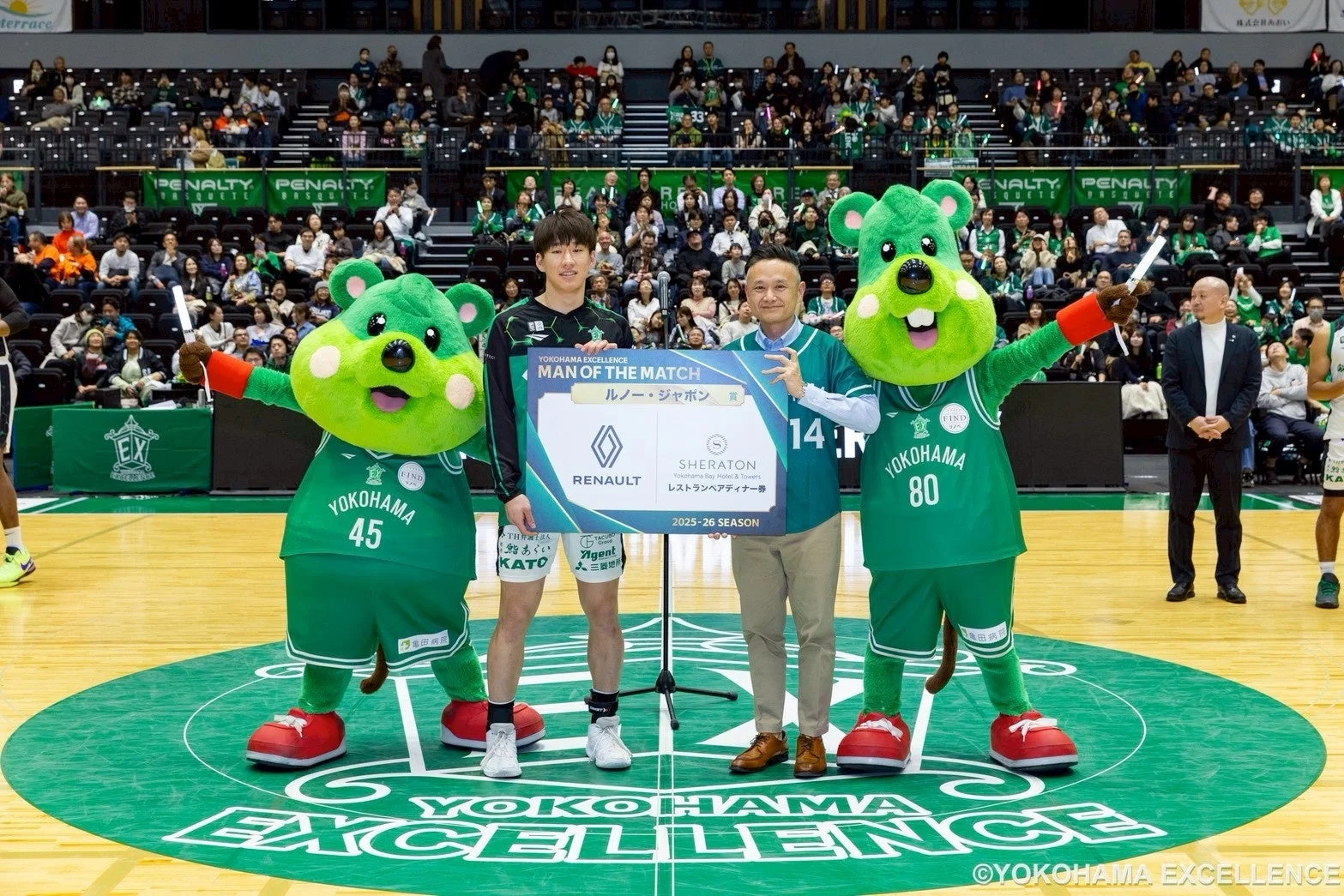 バスケットボールの試合後、選手と男性、そして2体の緑色のマスコットがコート上で「MAN OF THE MATCH」の表彰ボードを囲んでいます。ボードにはルノーとシェラトンのロゴが見え、背景には観客で埋まったアリーナが広がっています。