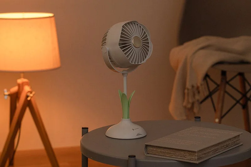 温かい間接照明が灯る部屋のサイドテーブルに、モダンなデザインの卓上扇風機と古い本が置かれている様子。落ち着いた雰囲気のリビングや寝室のインテリアを写した画像です。