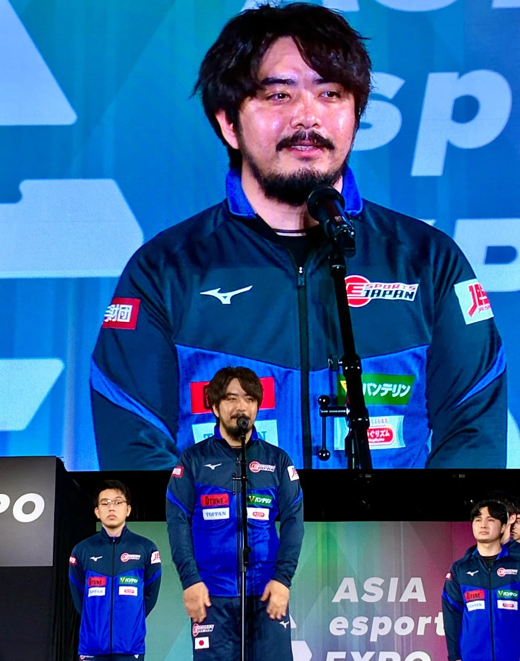 eスポーツイベント「ASIA esport EXPO」の壇上で、スポンサーロゴ入りのユニフォームを着た男性がマイクを使ってスピーチしている場面です。他の選手も壇上に立っています。