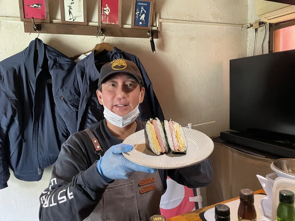 マスクとエプロンを着用した男性が、スパムと卵焼きを挟んだおにぎりを2つ載せた白い皿を差し出す