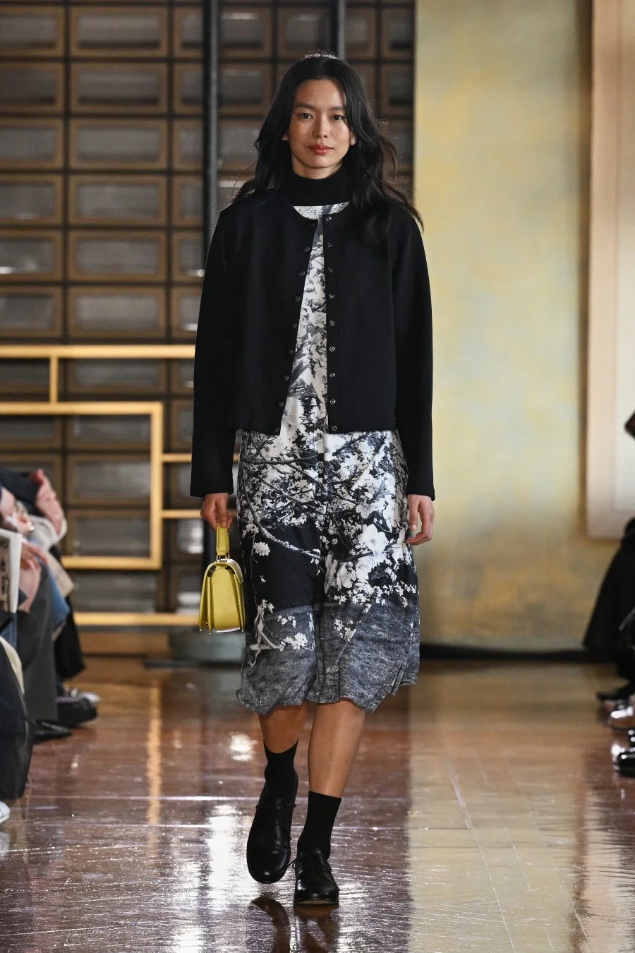 ファッションショーのランウェイを歩くアジア人モデル。黒いカーディガンと白黒の花柄ドレスを着用し、黄色のミニバッグをアクセントにしている。落ち着いた色合いの中に鮮やかな小物が映える、秋冬のコーディネート。