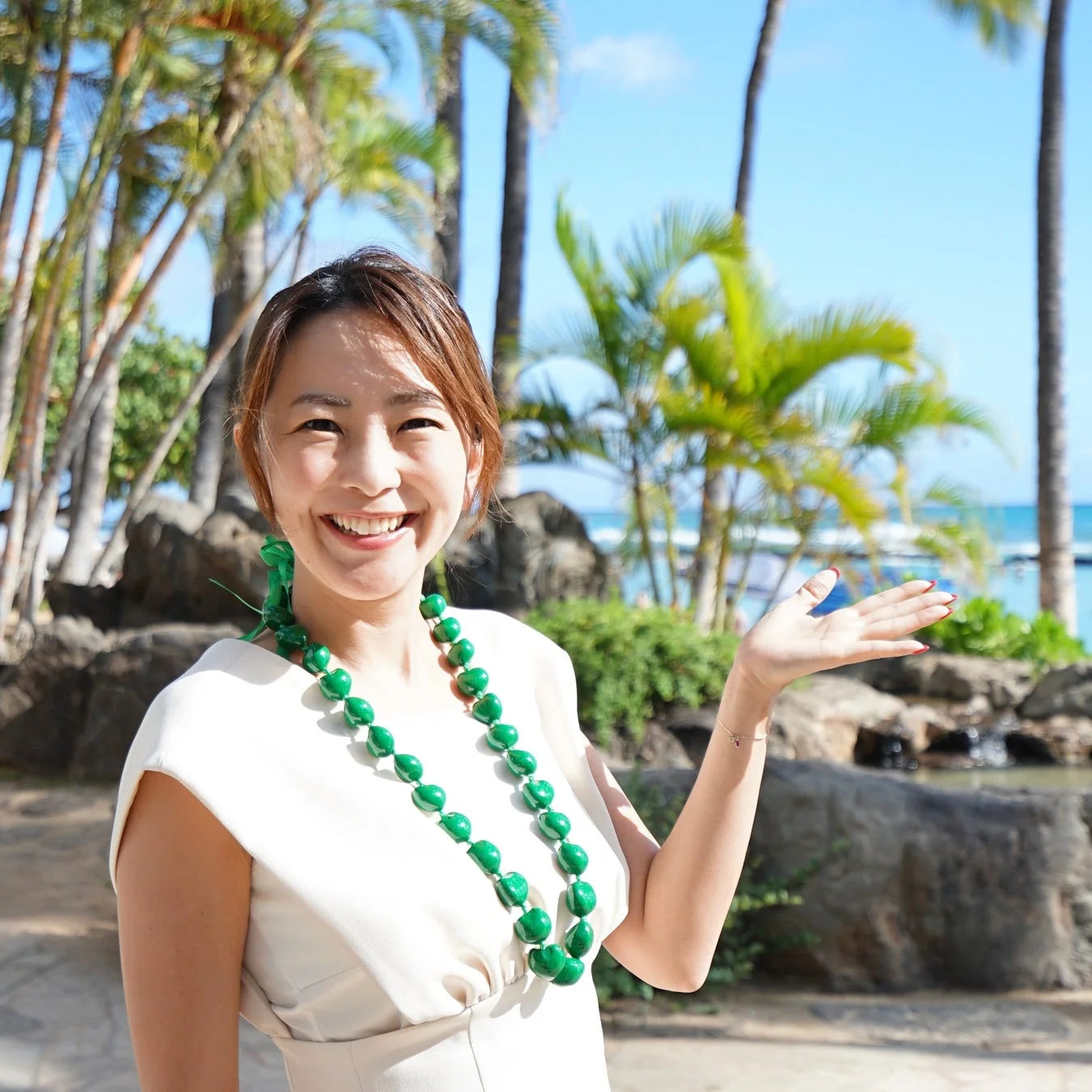 笑顔の女性が緑のレイを身につけ、手のひらを広げています。背景にはヤシの木と青い海が広がる、南国のリゾート地での楽しげな様子が捉えられています。