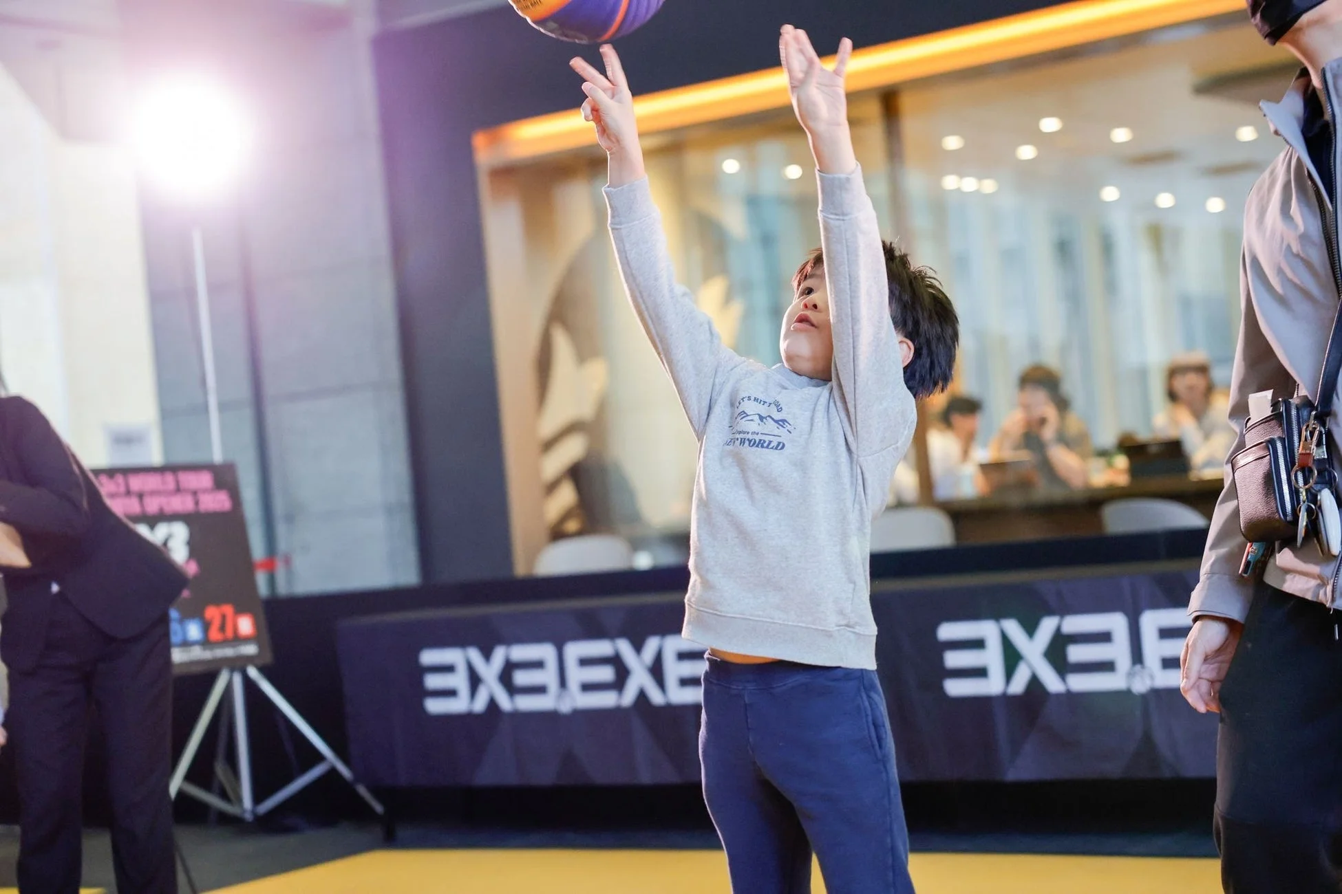 屋内イベント会場で、一人の子供がバスケットボールのシュートをしようとしている瞬間を捉えた写真