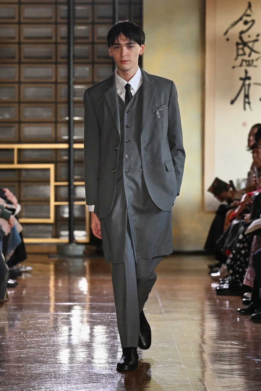 独特なデザインのグレーのスリーピーススーツを着用した男性モデルがランウェイを歩いている。背景には和風の障子のような壁と書道の掛け軸が見え、モダンと伝統が融合したファッションショーの様子を捉えている。