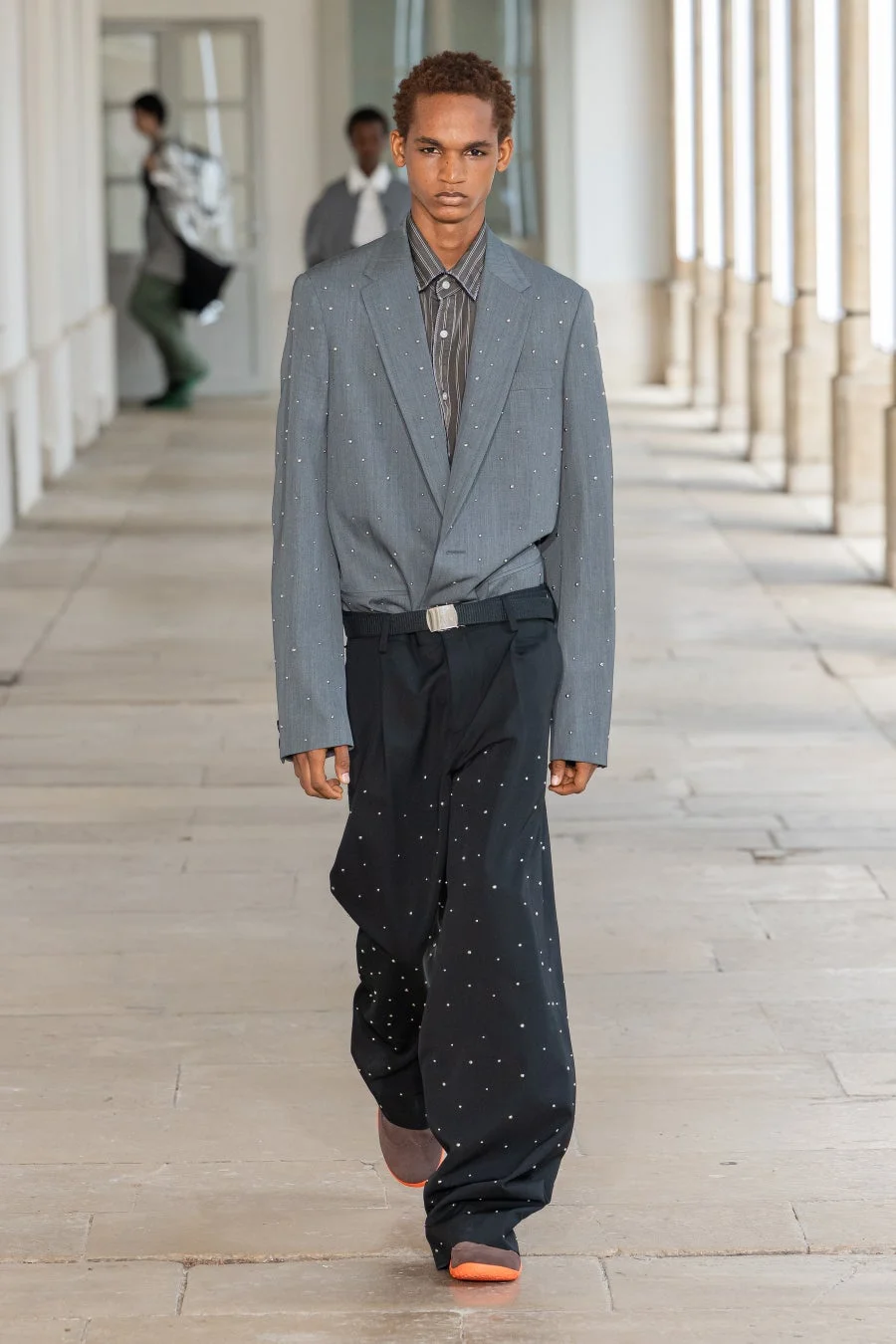 スタッズがあしらわれたグレーのブレザーと黒のワイドパンツを着用した男性モデル