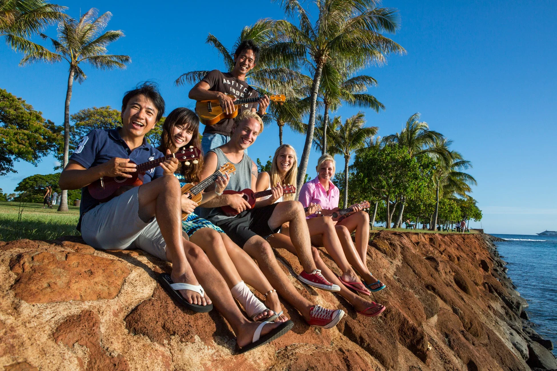 熱帯の青空の下、ヤシの木が並ぶ海岸で、6人の若者たちがウクレレを演奏しながら楽しそうに笑っている。海を背景に友情と音楽の喜びが伝わる瞬間を捉えた一枚。