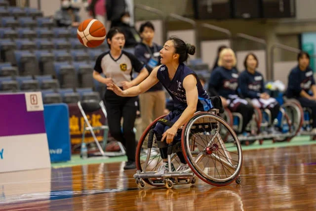 車椅子バスケットボールの試合中、女性選手が空中のボールに手を伸ばし、真剣な表情でプレイしている瞬間