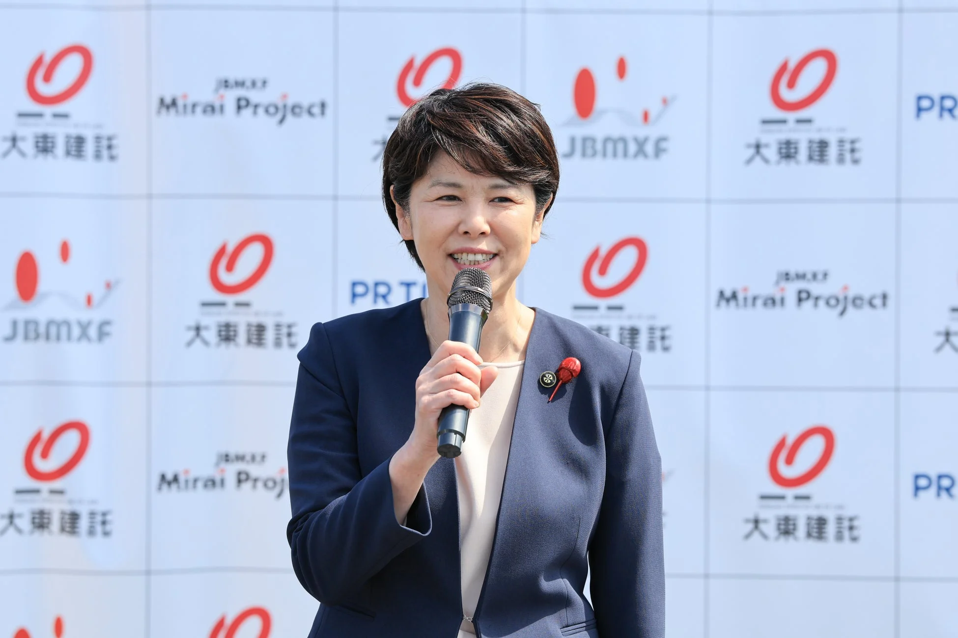 屋外イベントで、スーツ姿の女性がマイクを持って笑顔で話している