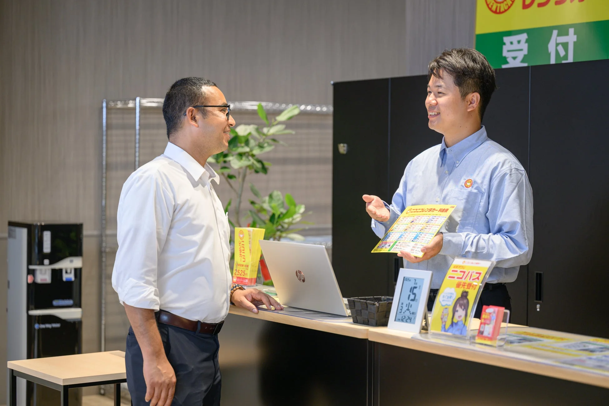 レンタカーの受付カウンターで、従業員が顧客に対して料金表などの資料を指し示しながら説明しています。二人の男性は笑顔で会話しており、丁寧な顧客対応が伺えるビジネスシーンです。