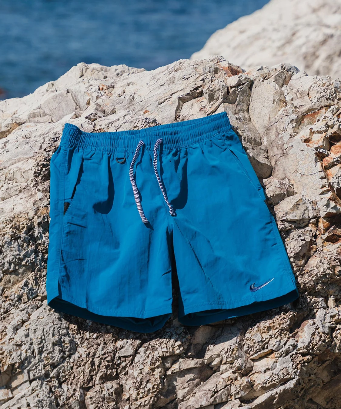 青いスイムショーツが岩の上に置かれており、背景には海が見える夏の風景です。