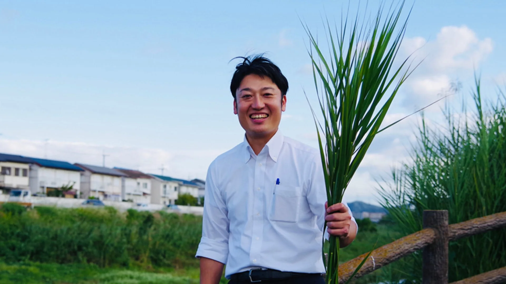 白いシャツを着た男性が、青空と住宅街を背景に、笑顔で大量の緑の草束を抱えています。