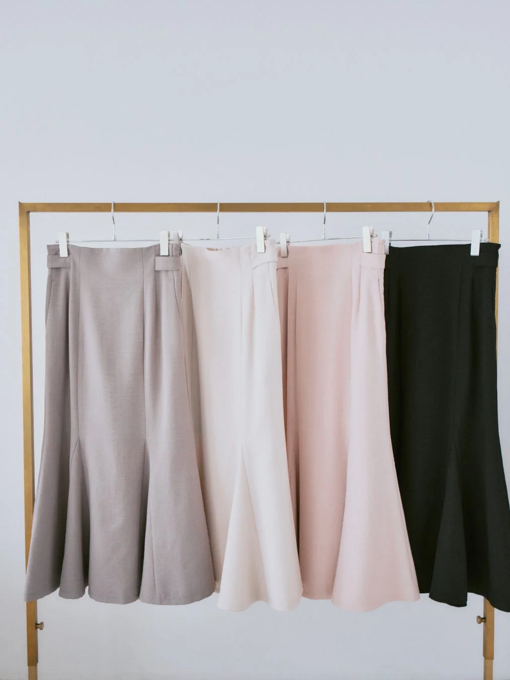 ゴールドのハンガーラックに、ベージュ、薄ピンク、濃いピンク、黒の4色のマーメイドスカートが並んで吊るされています。シンプルな背景で商品の色と形が際立っています。