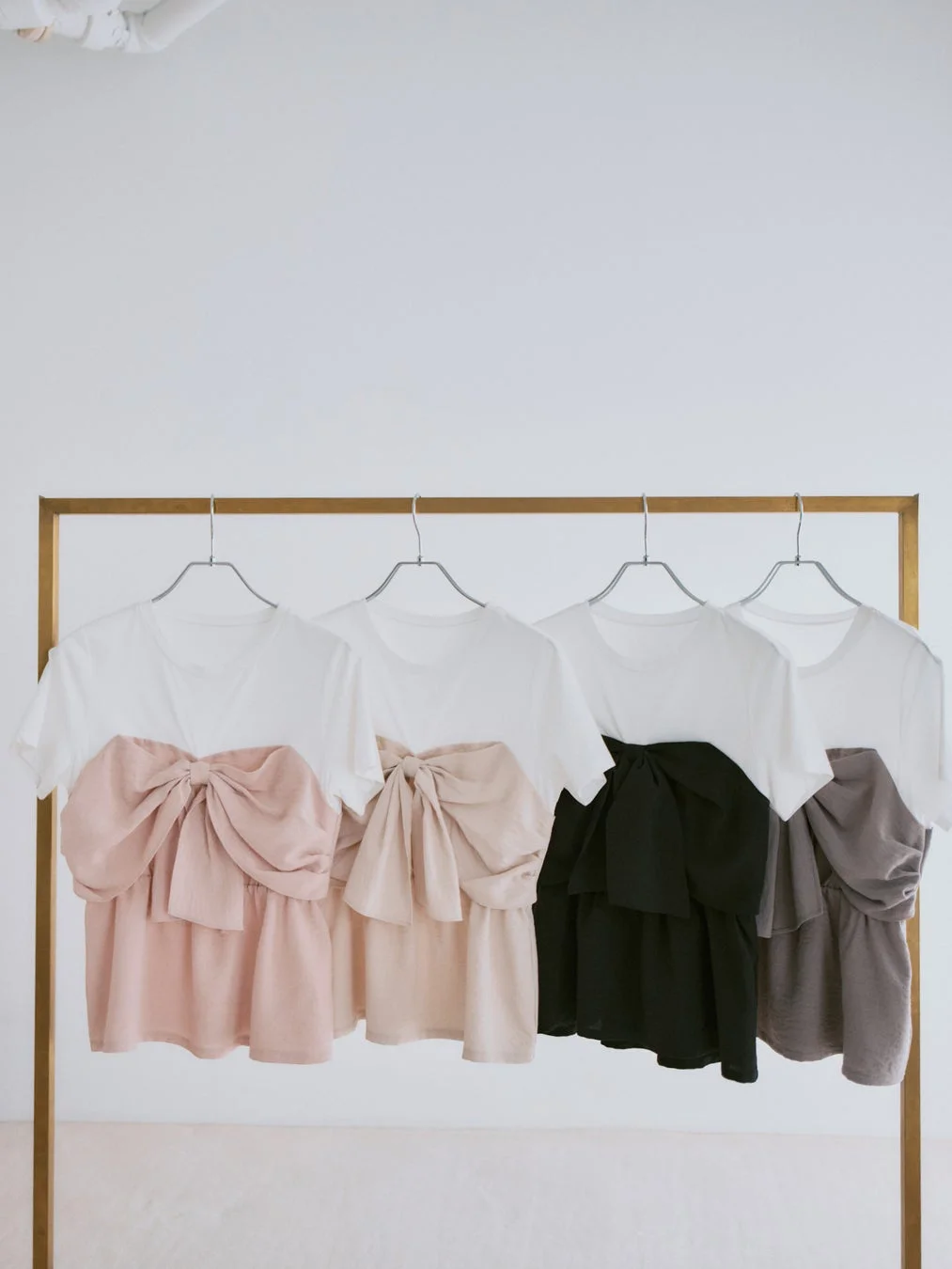 金色のハンガーラックに、白地に大きなリボンとフリルがあしらわれた半袖トップスが4色（ピンク、ベージュ、ブラック、グレー）で並べられています。ファッションアイテムの展示会のようです。