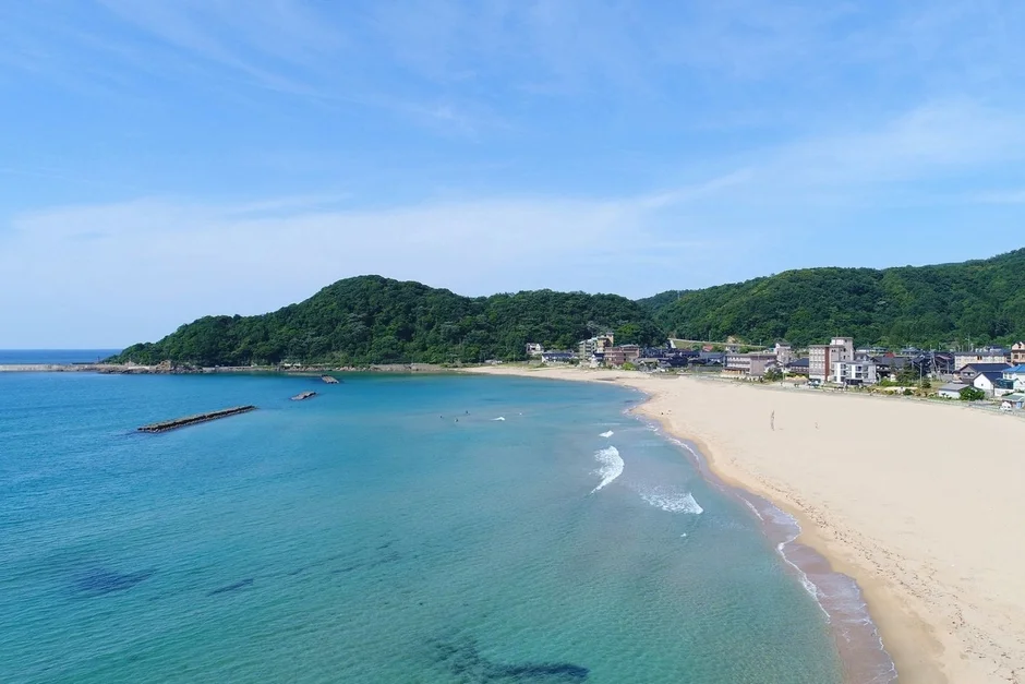 青い海と白い砂浜が広がる美しい海岸線の風景です。背後には緑豊かな山々が連なり、海岸沿いには小さな町が点在しています。