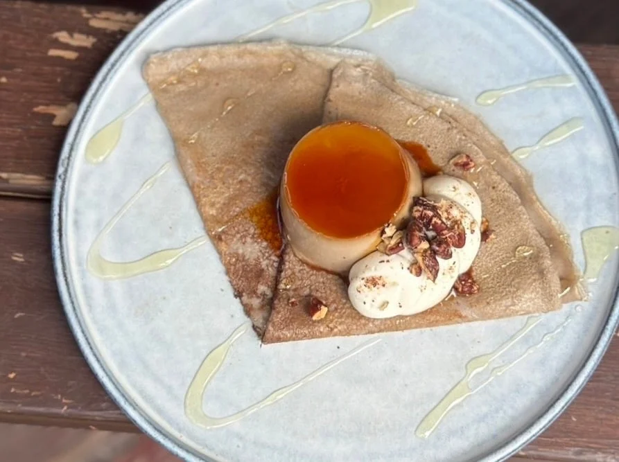 クレープの上にキャラメルソースのかかったプリンとホイップクリーム、砕いたナッツが添えられたデザート。皿にはソースで飾り付けがされている。