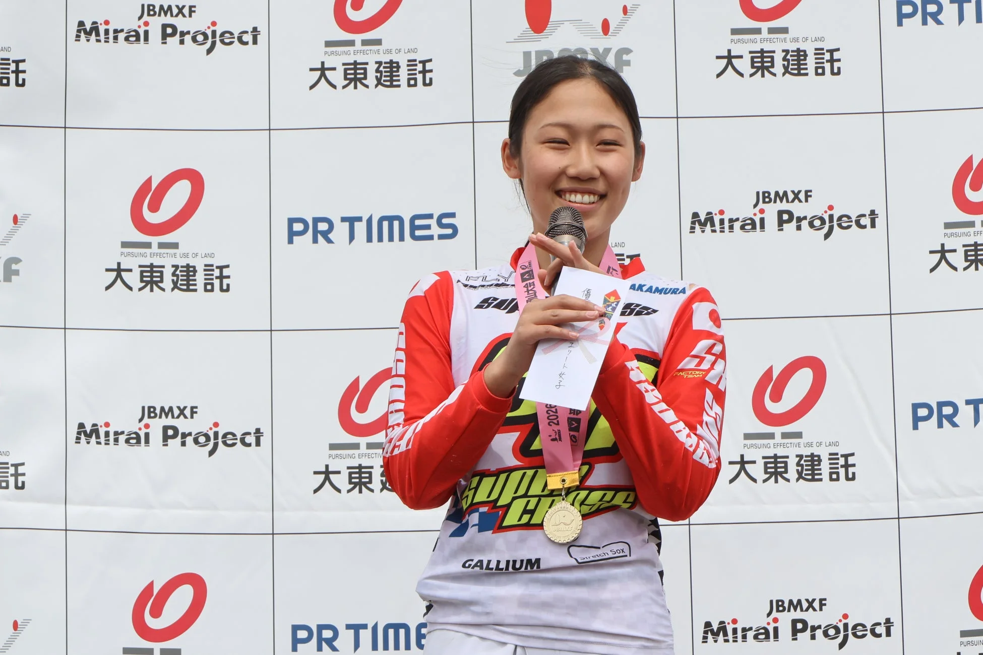 金メダルを首にかけ、マイクと封筒を持った若い女性アスリートが笑顔で表彰台に立っている