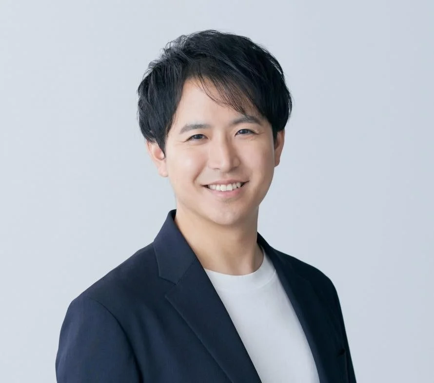 ダークなジャケットと白いTシャツを着用したアジア人男性が、カメラに向かって優しく微笑んでいる上半身のポートレート写真