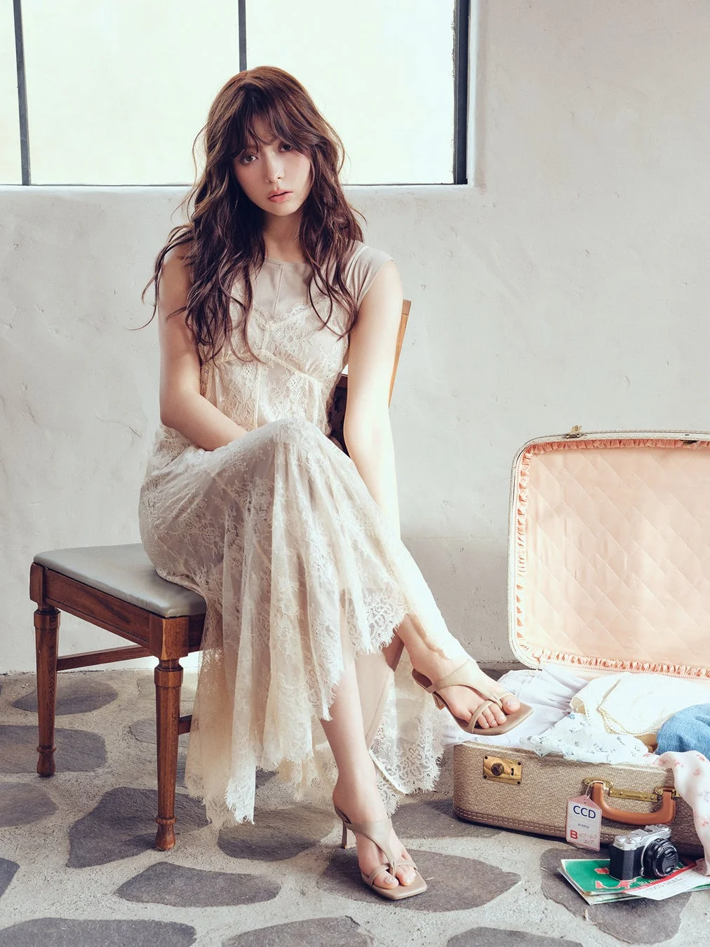 ベージュのレースワンピースを着た女性が椅子に座っている。横には開いた旅行トランク、カメラ、雑誌があり、旅の準備をしているような雰囲気の画像。