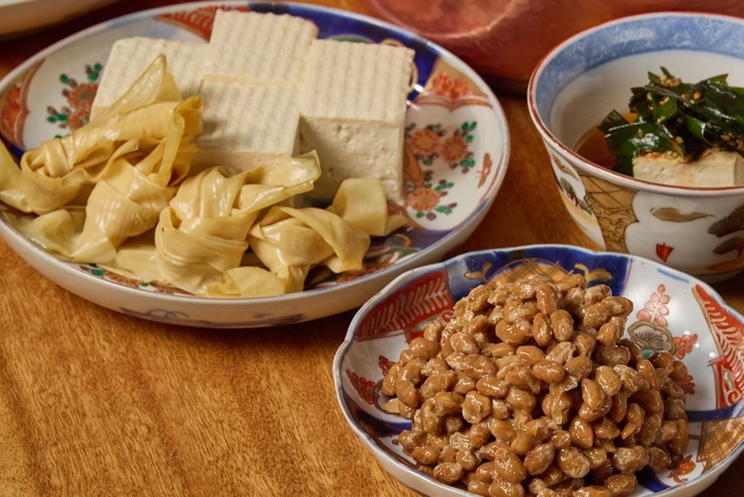 木目のテーブルに和食の皿が並べられ、手前には納豆、奥には湯葉と豆腐、ワカメと豆腐の小鉢がある