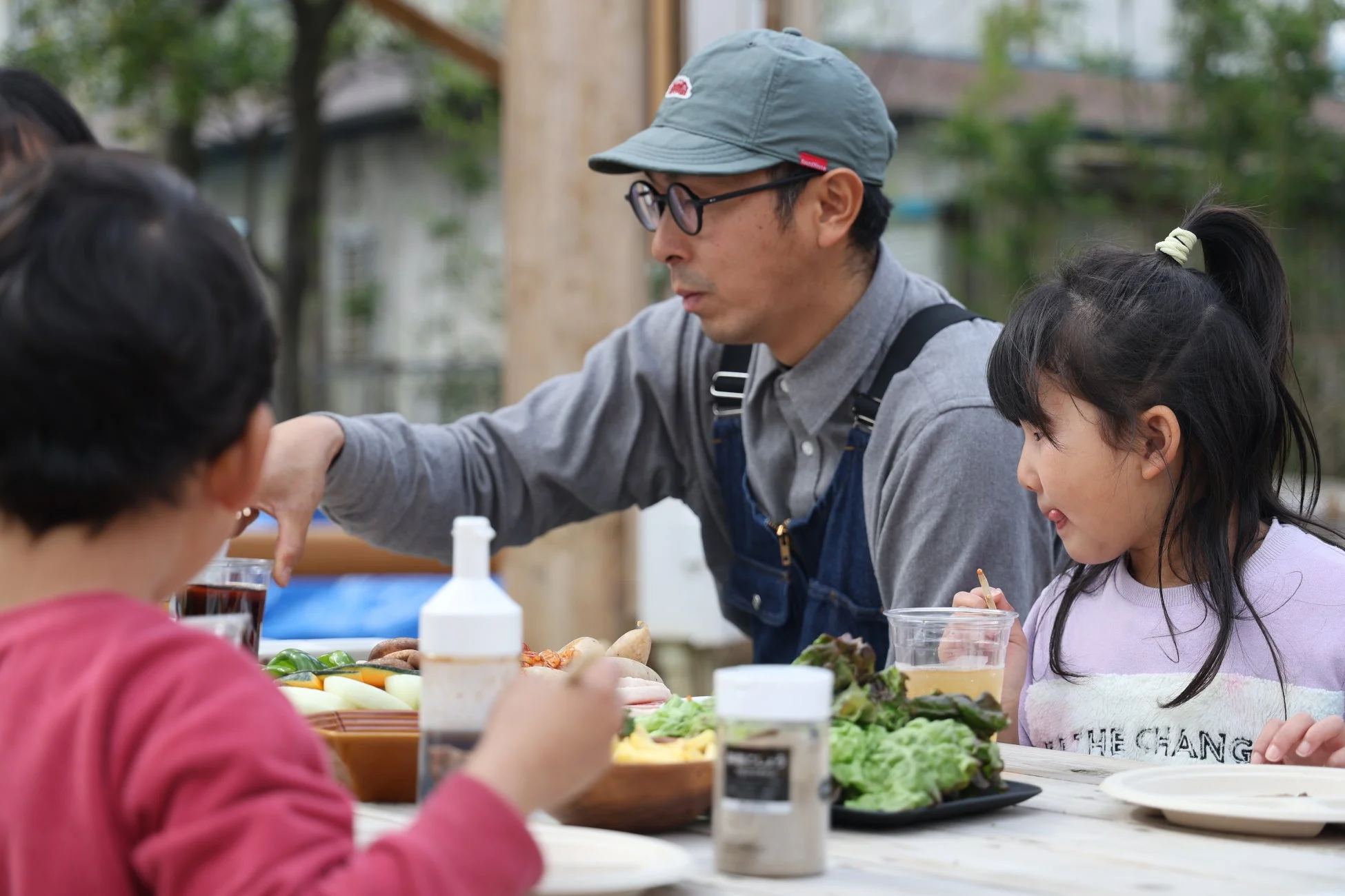 屋外で大人の男性と子供たちが食卓を囲み、新鮮な野菜や飲み物を楽しむ様子