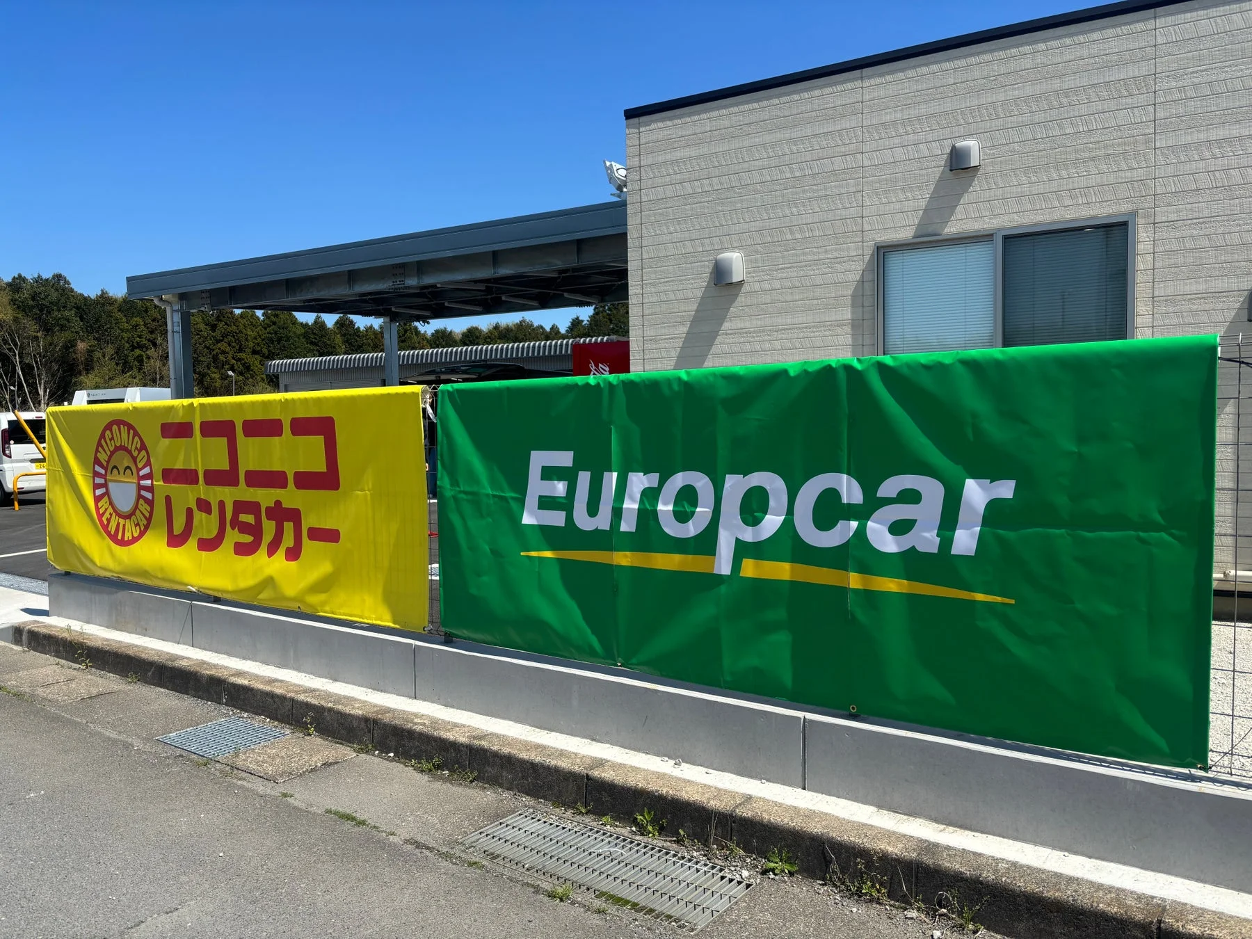 晴れた青空の下、ニコニコレンタカーとEuropcarの大きな看板が並んで設置されている屋外の様子です。レンタカー会社の施設の一部が写っており、ビジネス用途の広告が目立ちます。