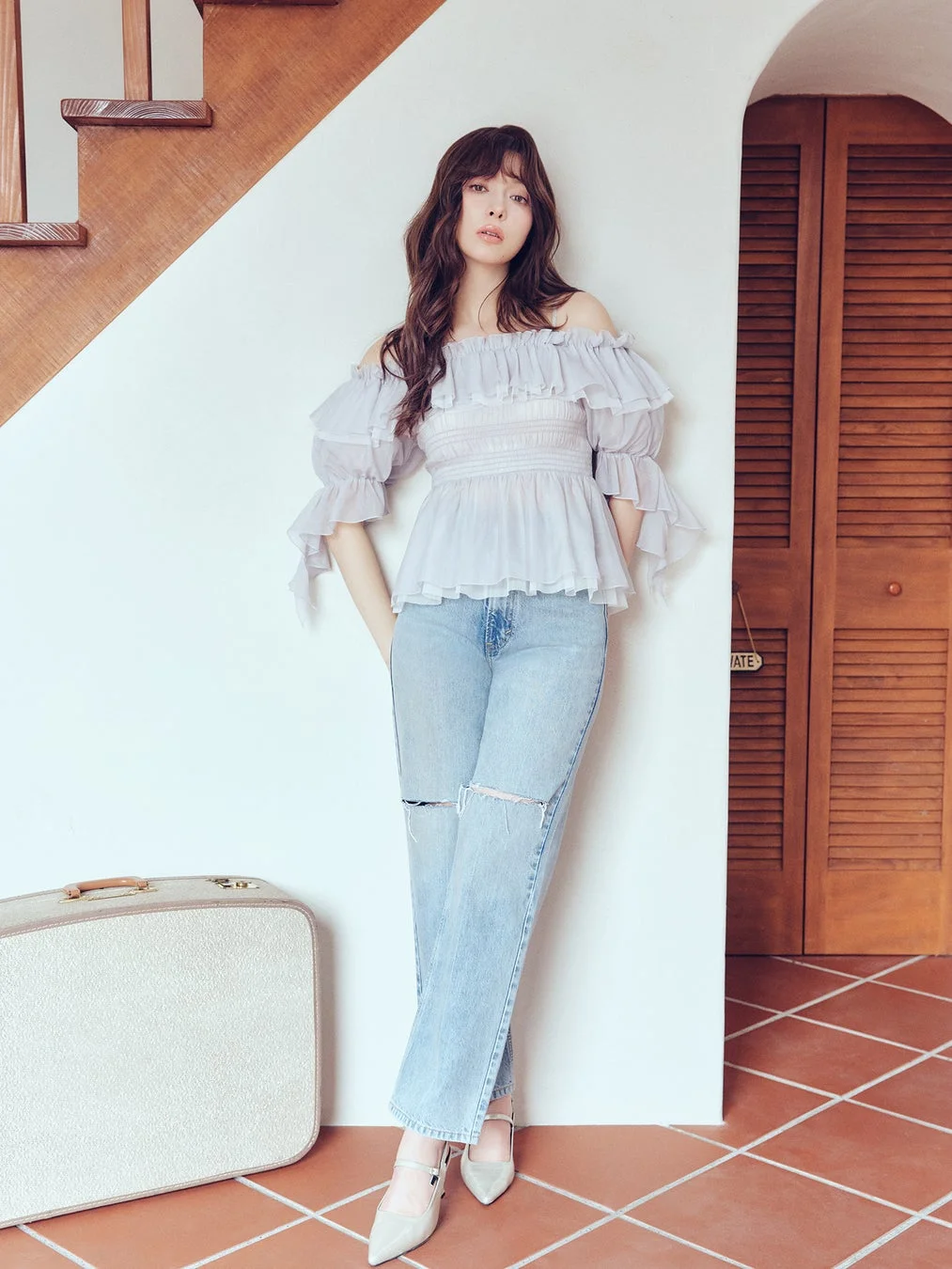 淡い水色のオフショルダートップスとダメージジーンズを着用した女性が、階段とスーツケースのある屋内でポーズをとっています。