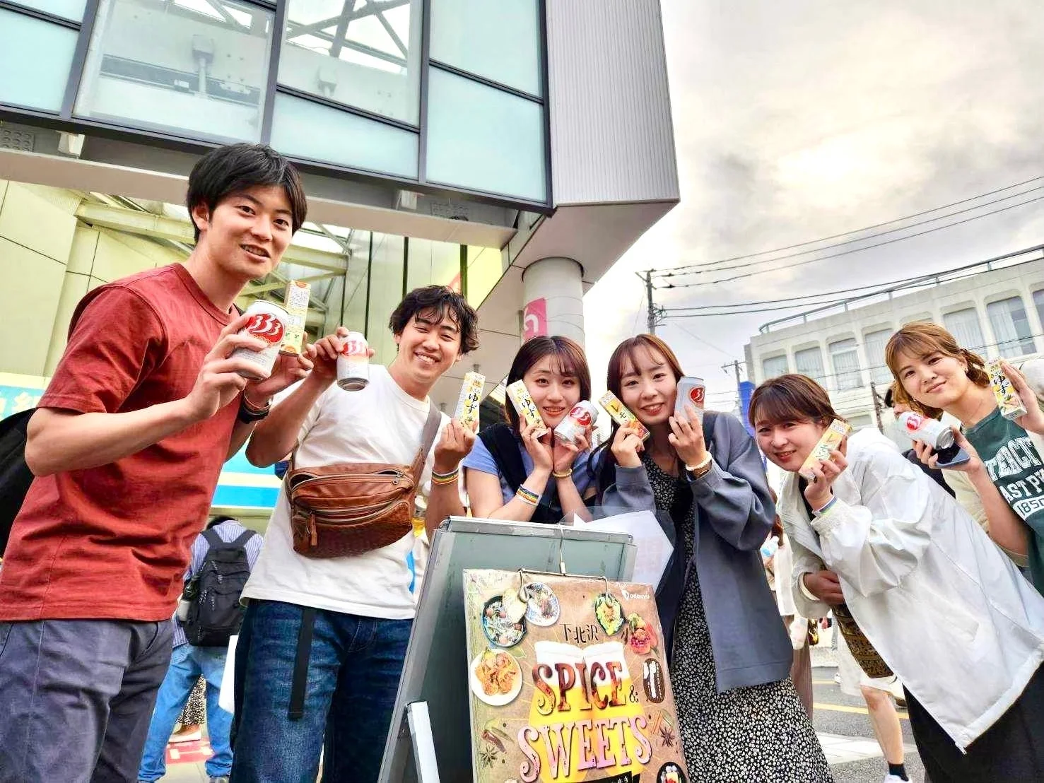 屋外で笑顔の男女グループが、缶飲料やゆず・レモン味の小箱を手に持っています。近くには「SPICE & SWEETS」と書かれた看板があり、何らかのプロモーション活動をしている様子です。
