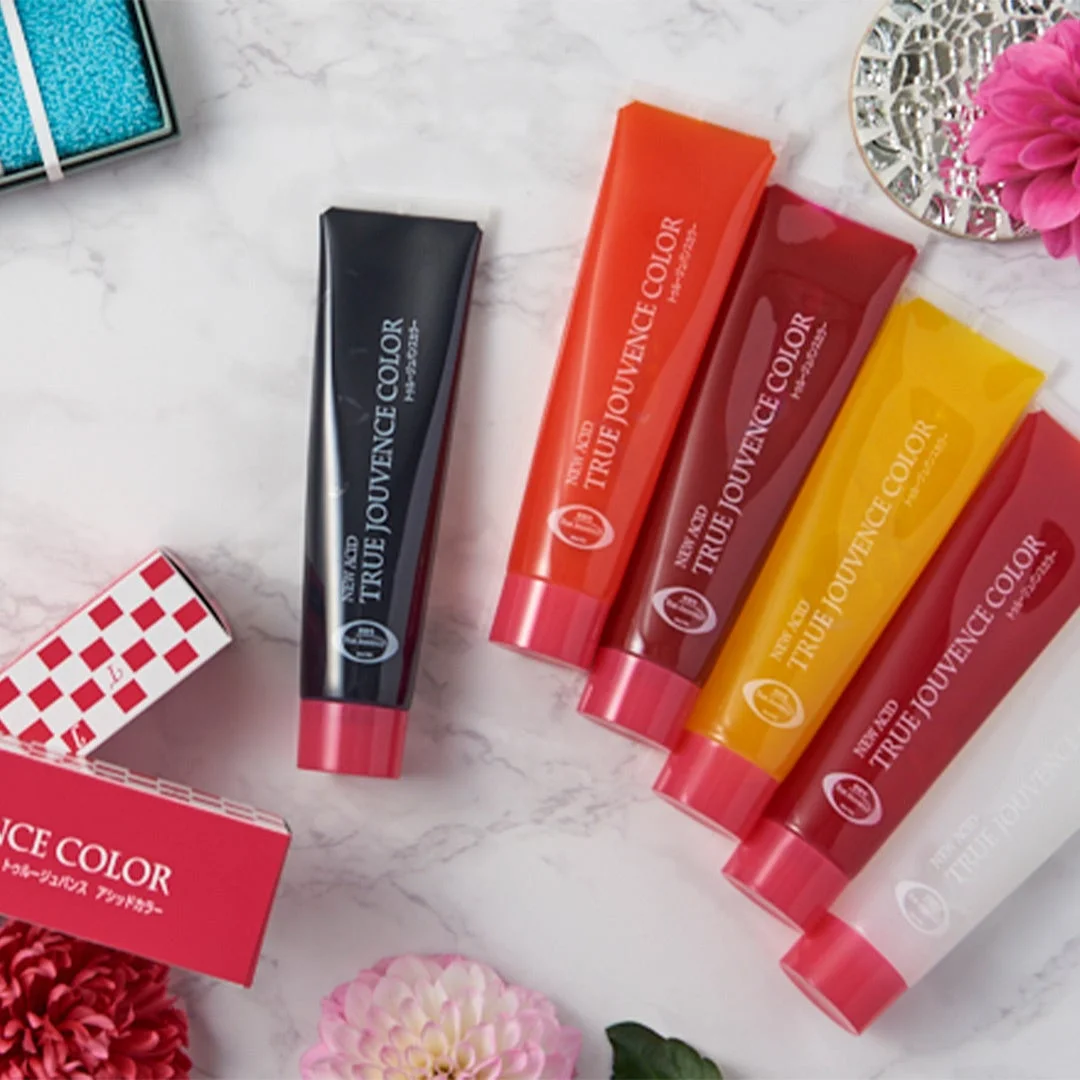 大理石柄の背景に、黒、オレンジ、赤、黄、ピンク、白など様々な色の「TRUE JOUVENCE COLOR」と書かれたヘアカラートリートメントのチューブが並べられています。一部はパッケージ箱に入っており、周囲にはピンクや赤の花が飾られ、明るく華やかな美容製品のイメージを伝えています。