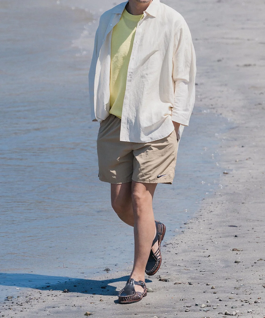 ビーチまたは水辺を歩く男性の全身が写っています。オフホワイトのシャツを羽織り、黄色のTシャツとベージュのショートパンツ、そして特徴的なサンダルを着用しており、夏のカジュアルな装いです。