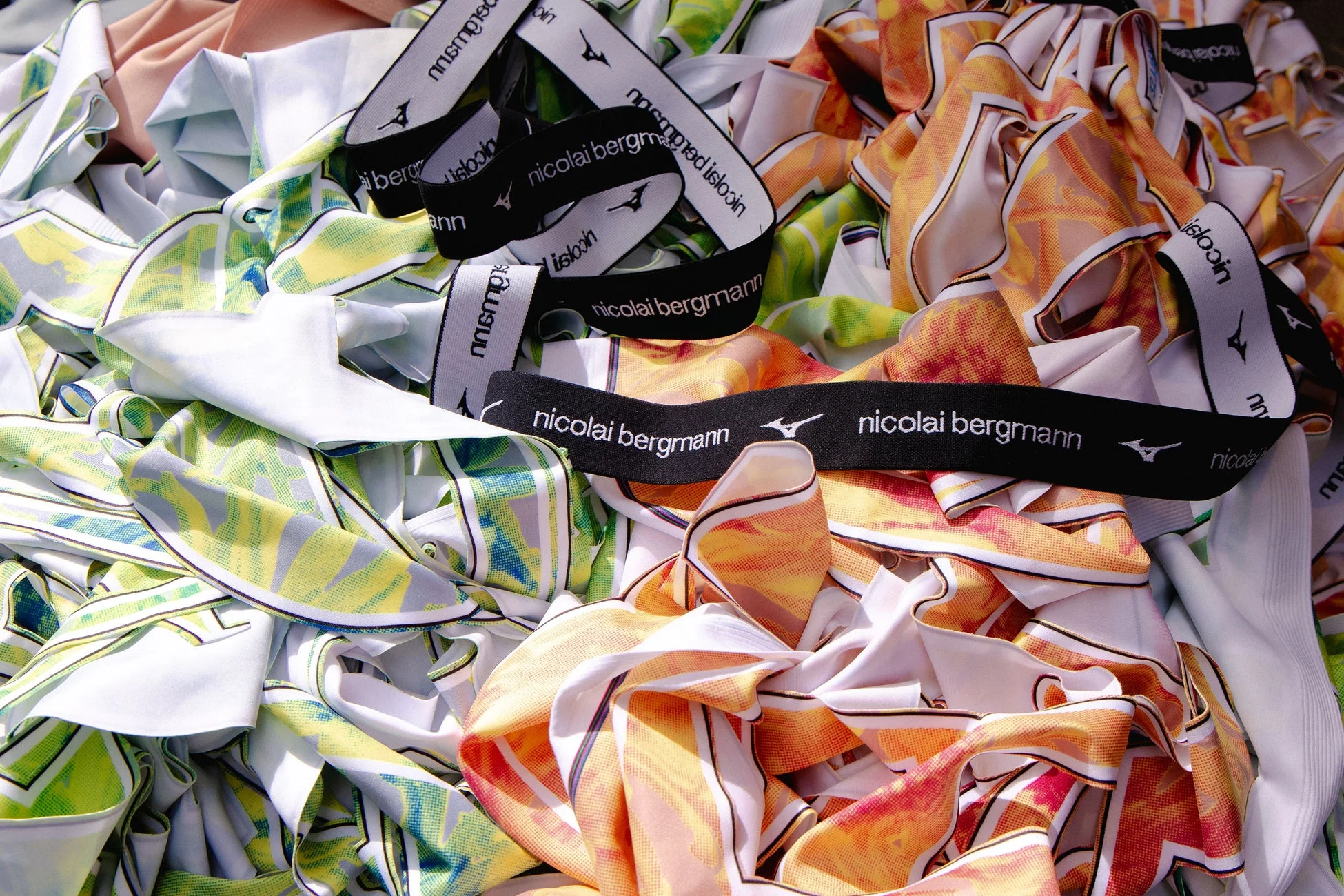「nicolai bergmann」のロゴとミズノのロゴが入ったリボンが散らばる、緑と黄色の幾何学模様、およびオレンジと赤の抽象的な模様が特徴的な生地やスカーフの山。カラフルなテキスタイルが目を引く景観です。