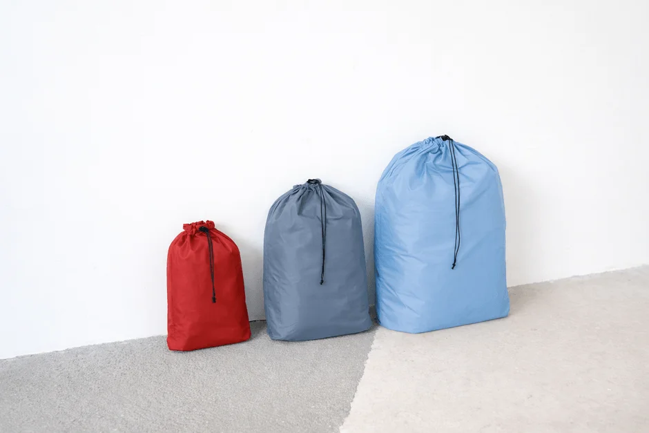 白い壁を背景に、赤、グレー、水色の異なるサイズの巾着袋が3つ並んでいます。収納や整理に便利なシンプルなデザインで、旅行用品やランドリーバッグとしても使えそうです。