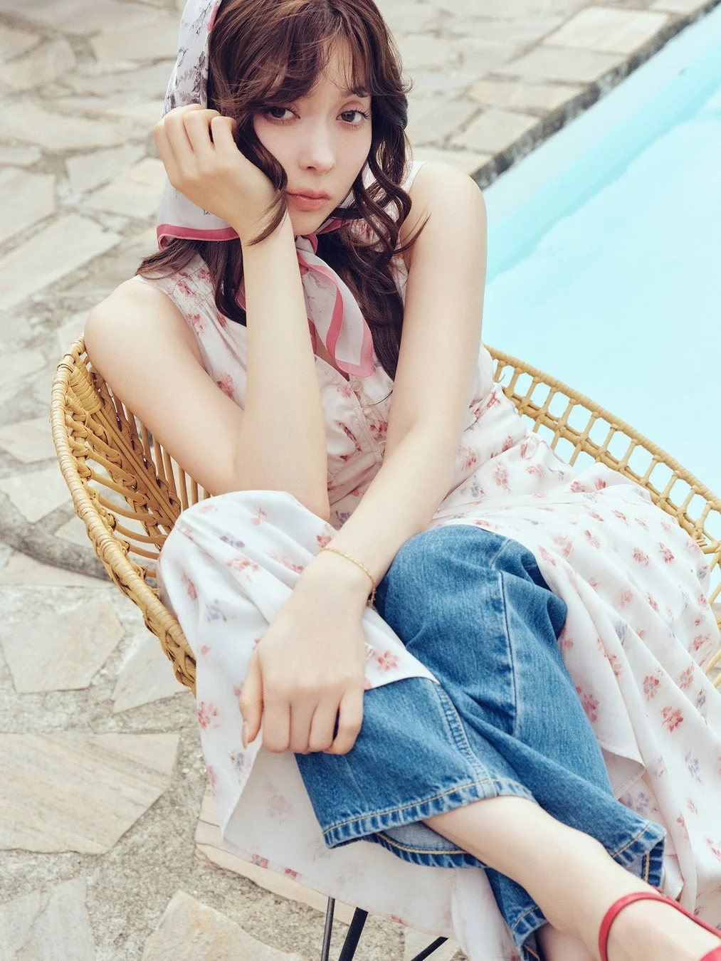 プールサイドのラタンチェアに座る女性のポートレート。花柄のワンピースとデニムを重ね着し、スカーフを巻いて物憂げな表情でカメラを見つめている。