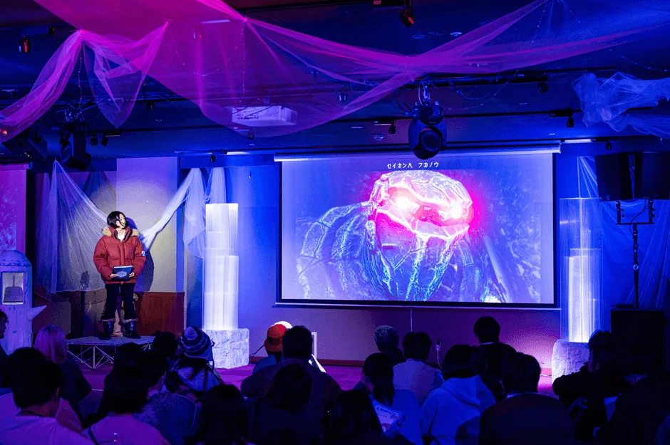 青と紫の照明で演出された会場で、観客がスクリーンを見つめている様子