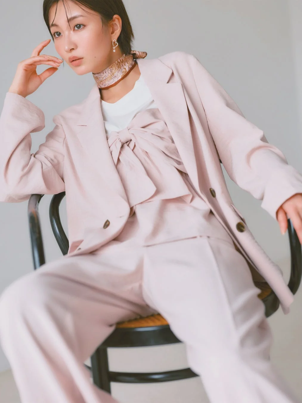 淡いピンクのスーツを着た女性が椅子に座っているポートレート。ジャケットとパンツのセットアップに、首元には柄物のスカーフを合わせ、上品で洗練されたファッションスタイルを披露している。