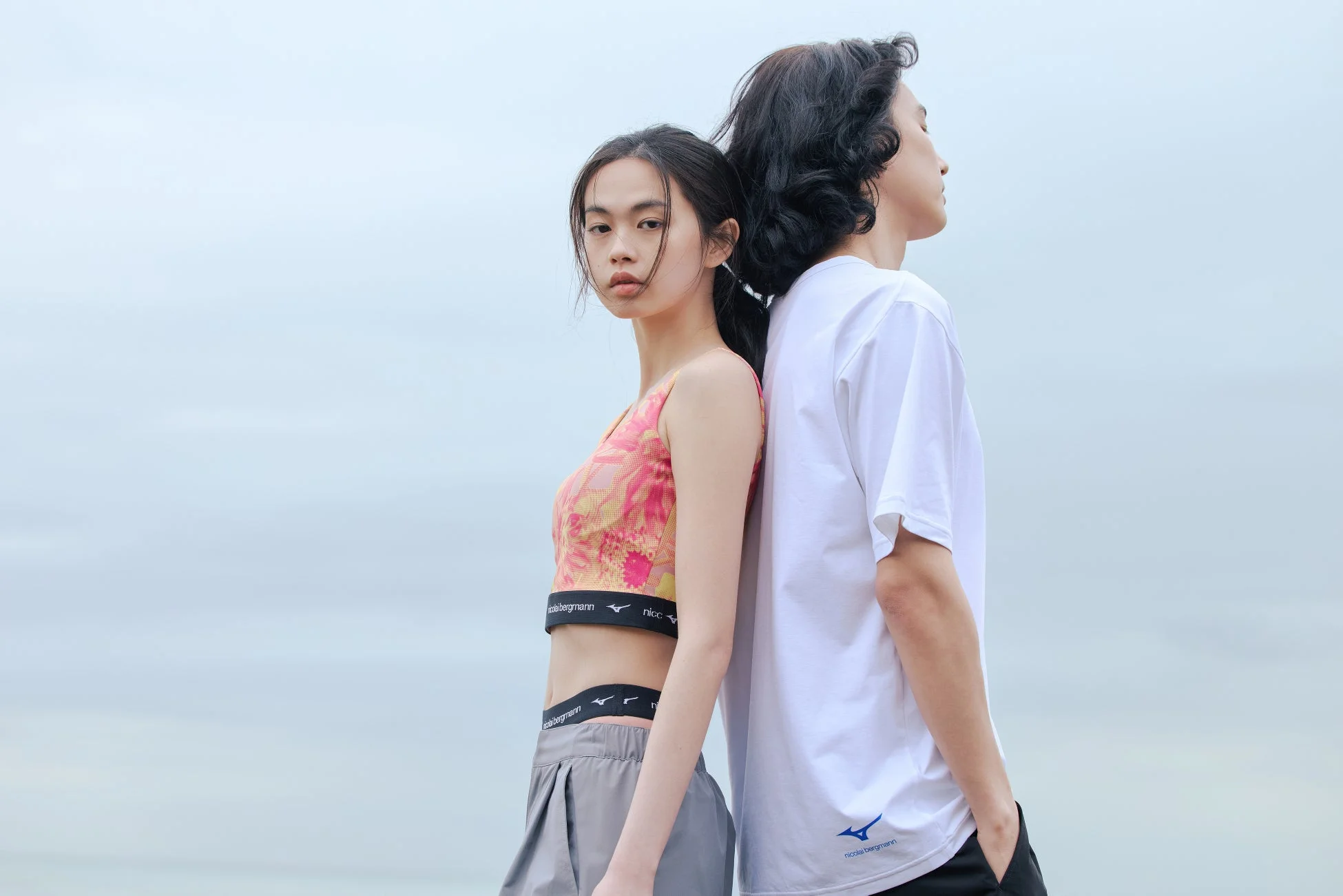 淡い空を背景に、男女二人が背中合わせに立っている。女性はカメラを見ており、男性は横を向いている。二人はミズノとニコライバーグマンのブランドのスポーツウェアを着用している。