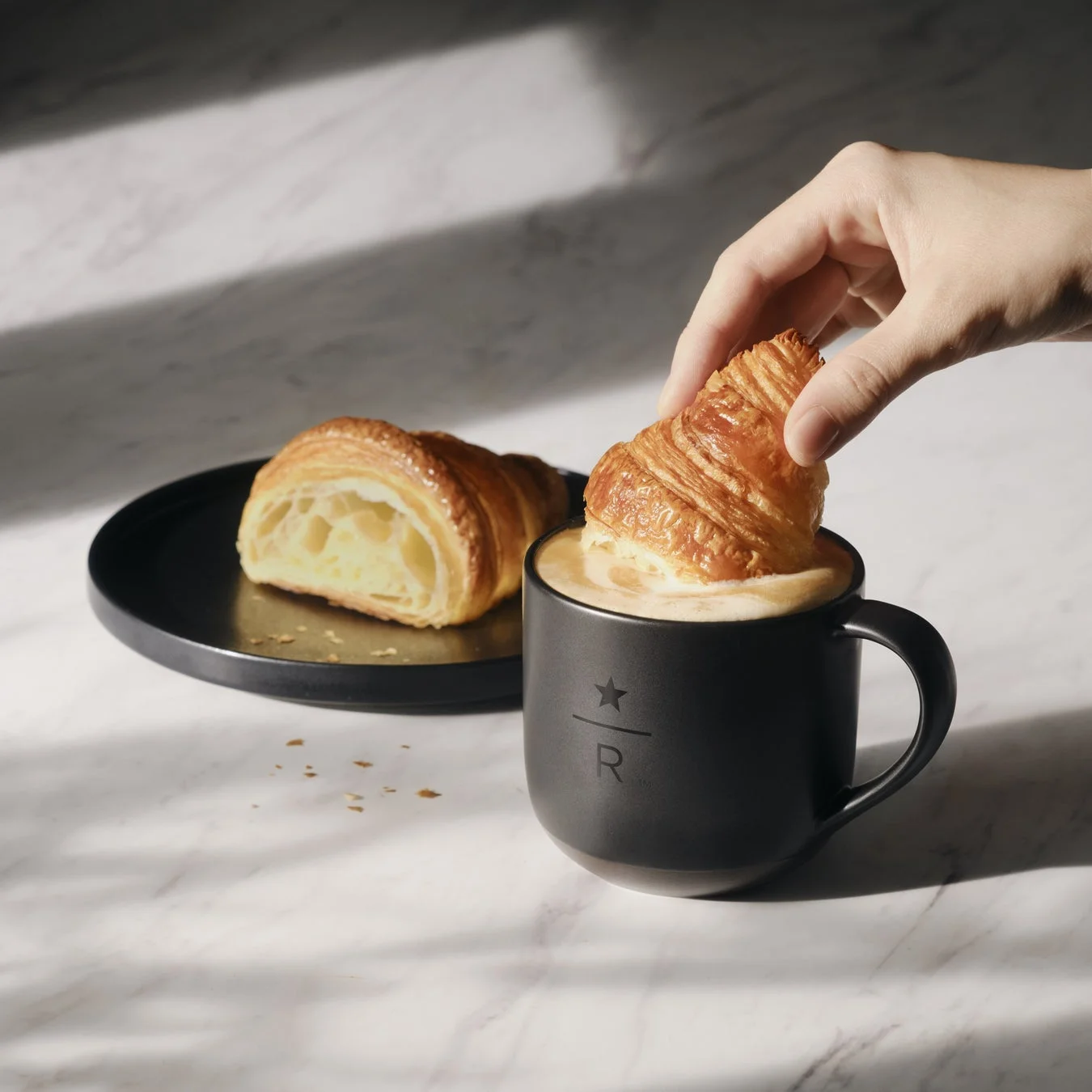 温かいコーヒーが入った黒いマグカップにクロワッサンを浸す手が写る、朝食の情景。