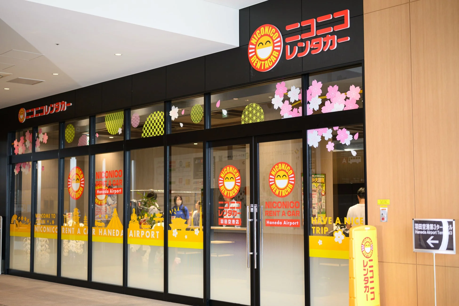 羽田空港にあるニコニコレンタカーの店舗です。桜や和柄の装飾が施されたガラス窓が特徴で、日本への訪問者を歓迎するメッセージが書かれています。