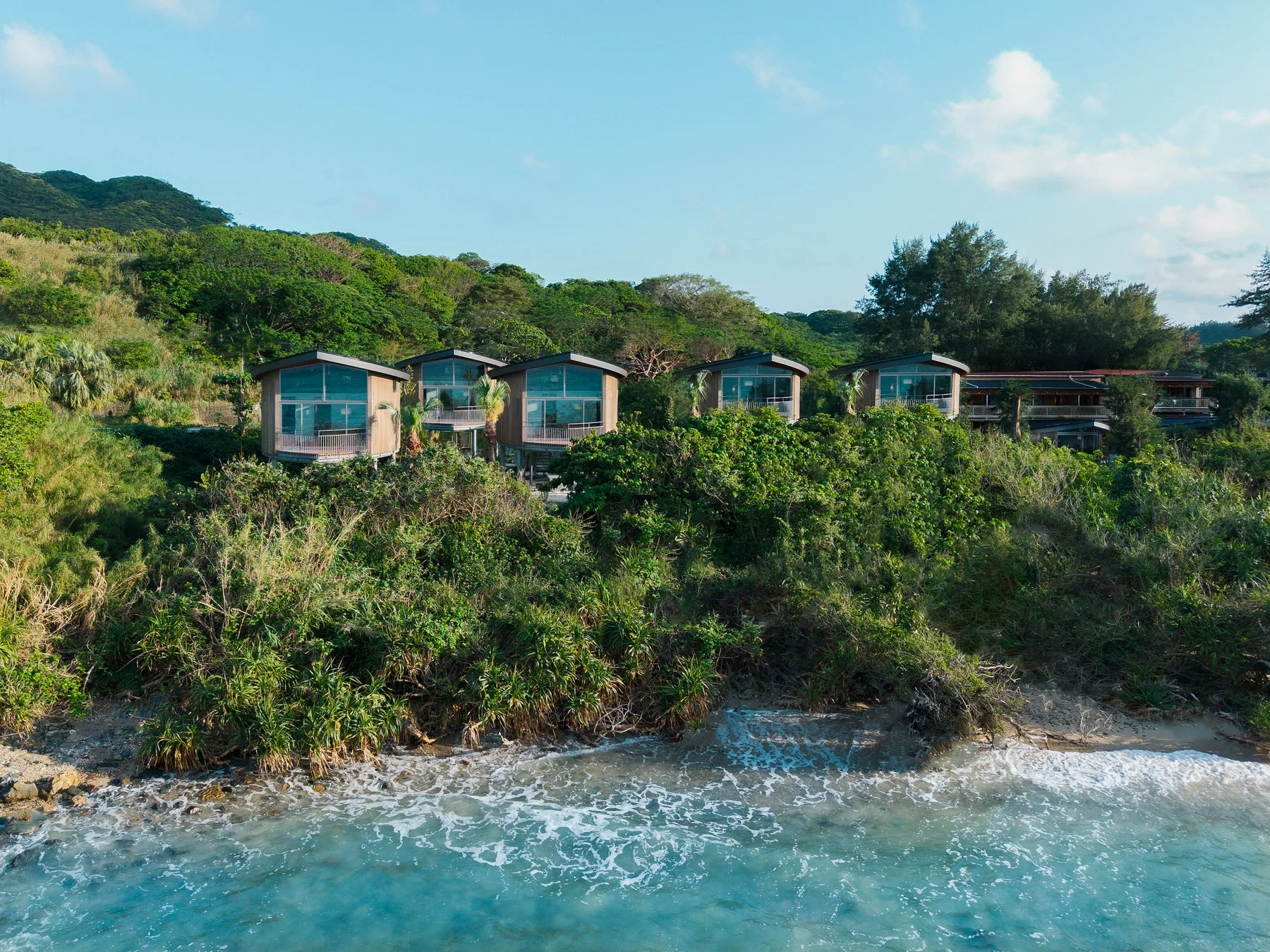 青い海と緑豊かな丘の斜面に並ぶモダンなヴィラ群の空撮