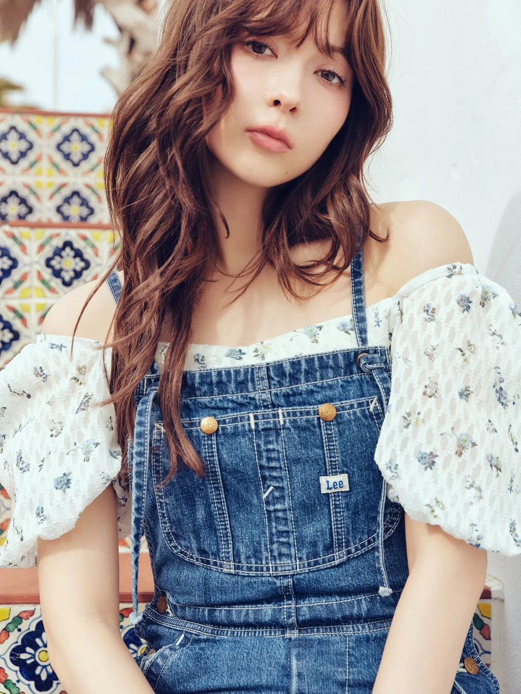 デニムのオーバーオールと花柄のオフショルダーブラウスを着た若い女性が、装飾的なタイル張りの階段に座ってポーズをとっているポートレート。