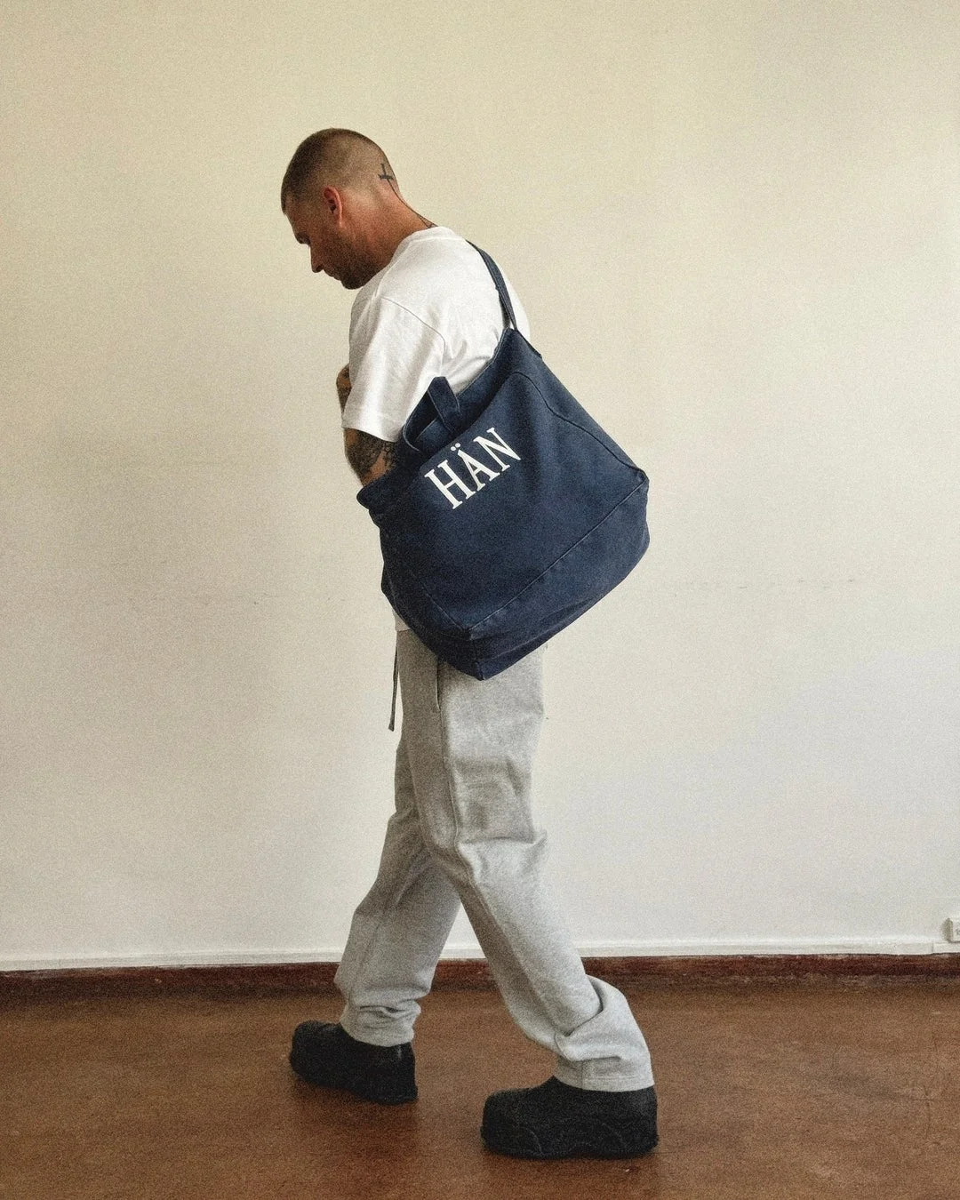 HÄNのロゴ入りトートバッグとスウェットパンツを着用した男性