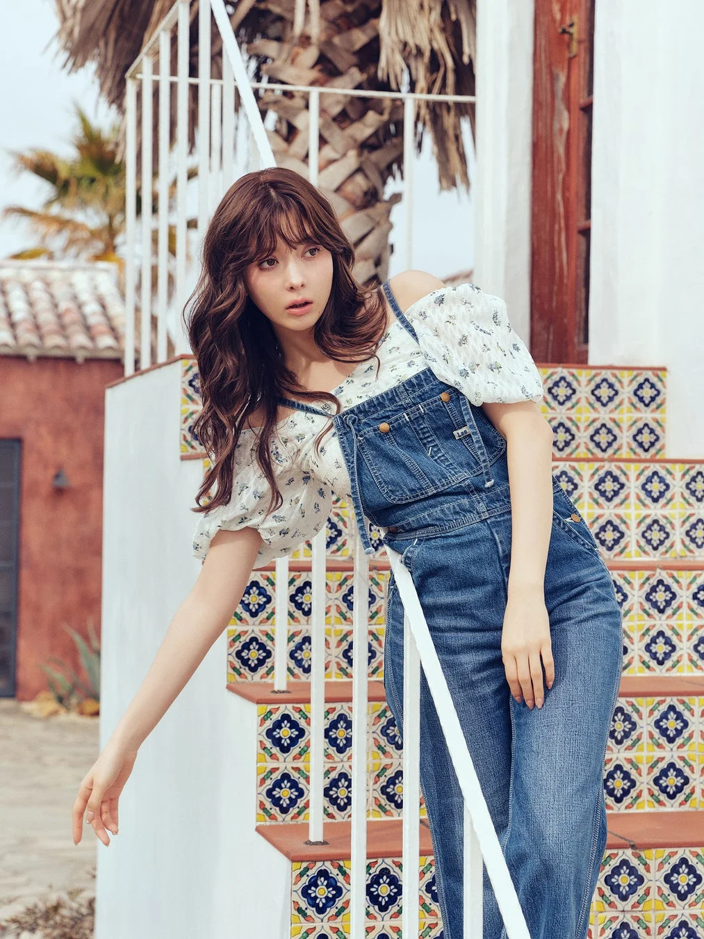 デニムのオーバーオールと花柄のオフショルダートップスを着た女性が、カラフルなタイル張りの階段にポーズをとっています。