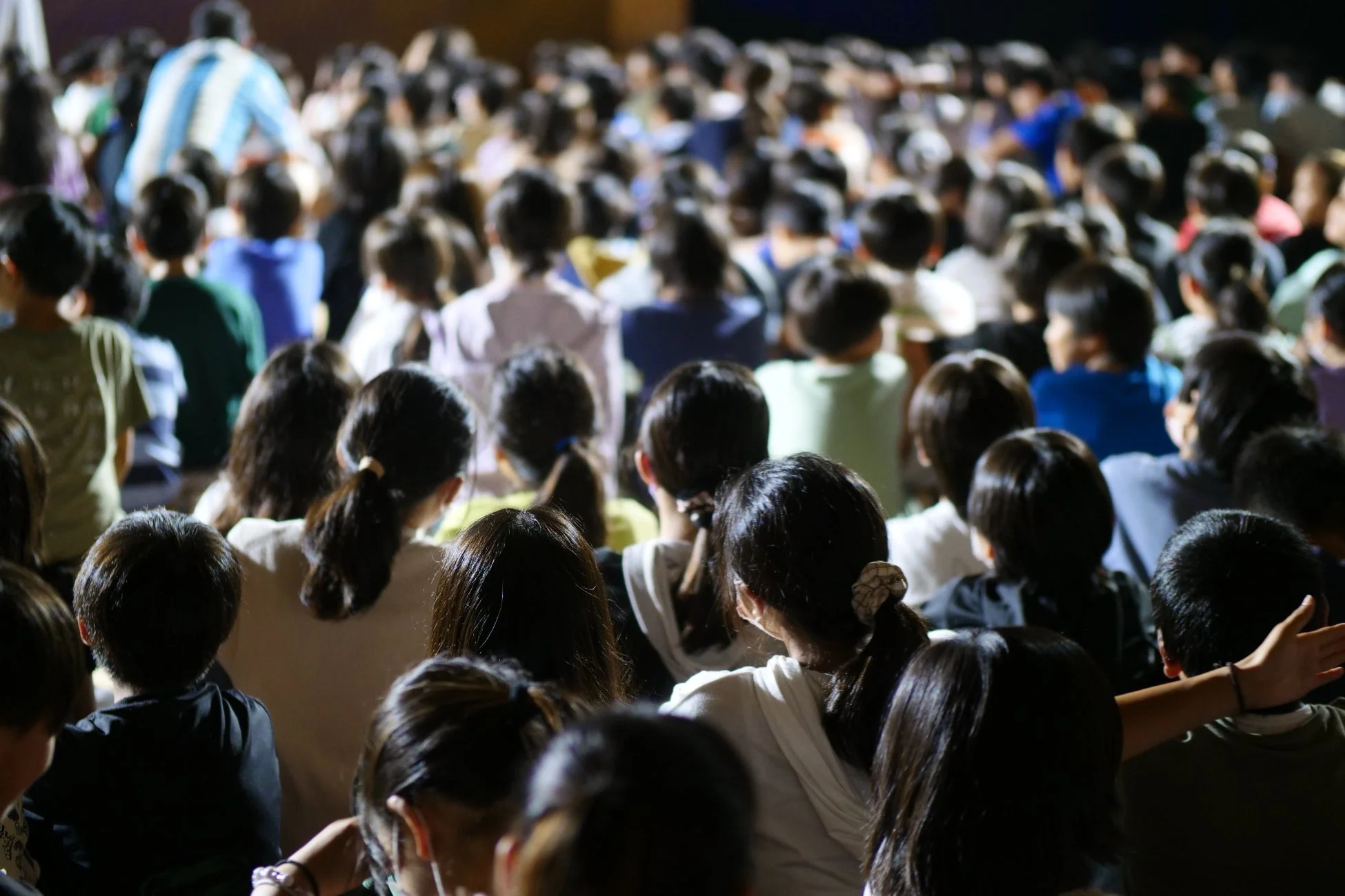 多くの人々、主に子供たちが、講堂かホールのような場所で前に向かって座っている様子