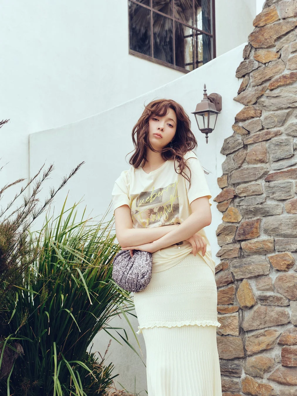 クリーム色のTシャツとロングスカートを着た若い女性が、屋外でポーズをとっています。