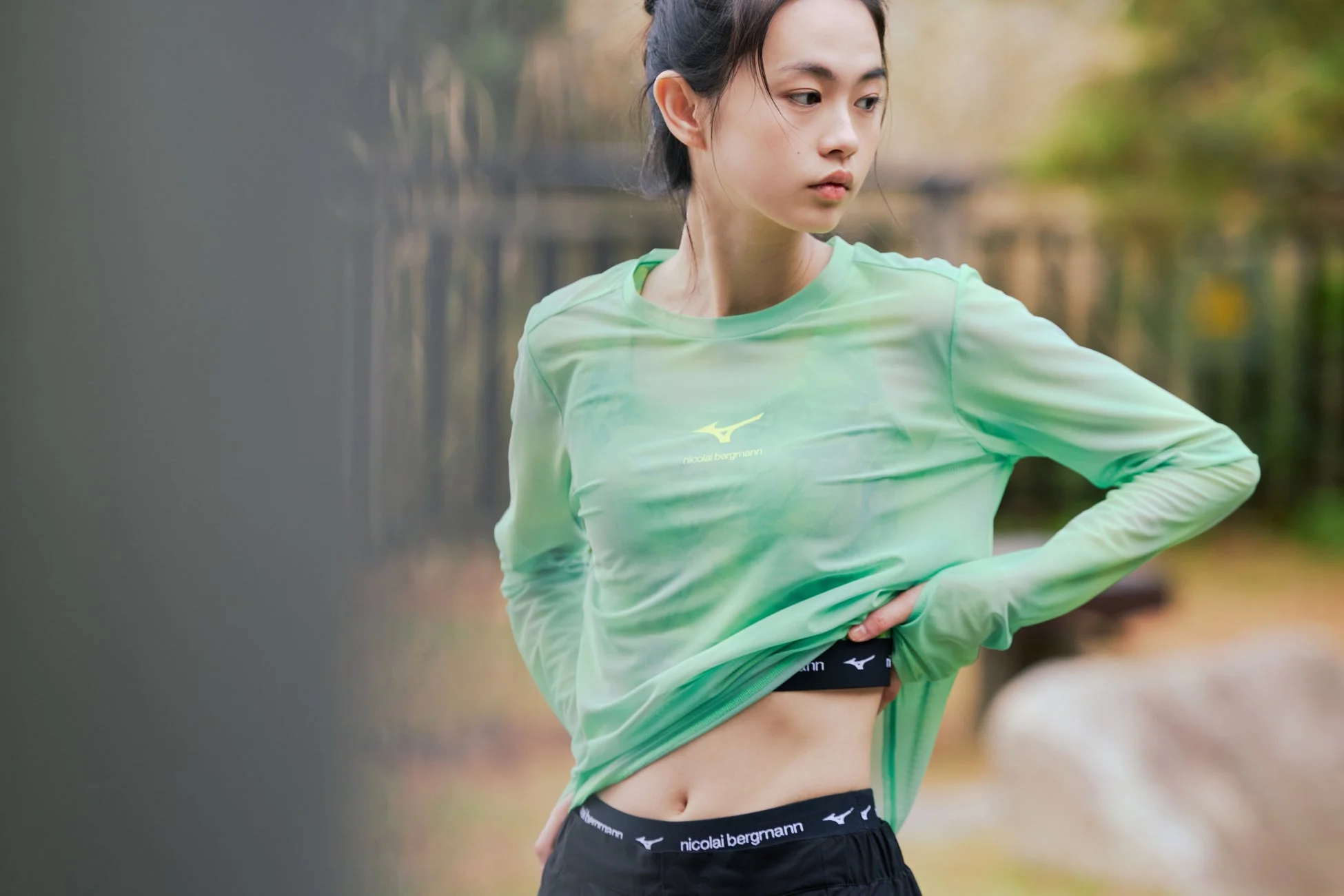 ミントグリーンの長袖スポーツウェアと黒いショートパンツを着用した若い女性が屋外に立っています。服にはミズノとニコライバーグマンのロゴが見られ、アクティブなライフスタイルを表現しています。