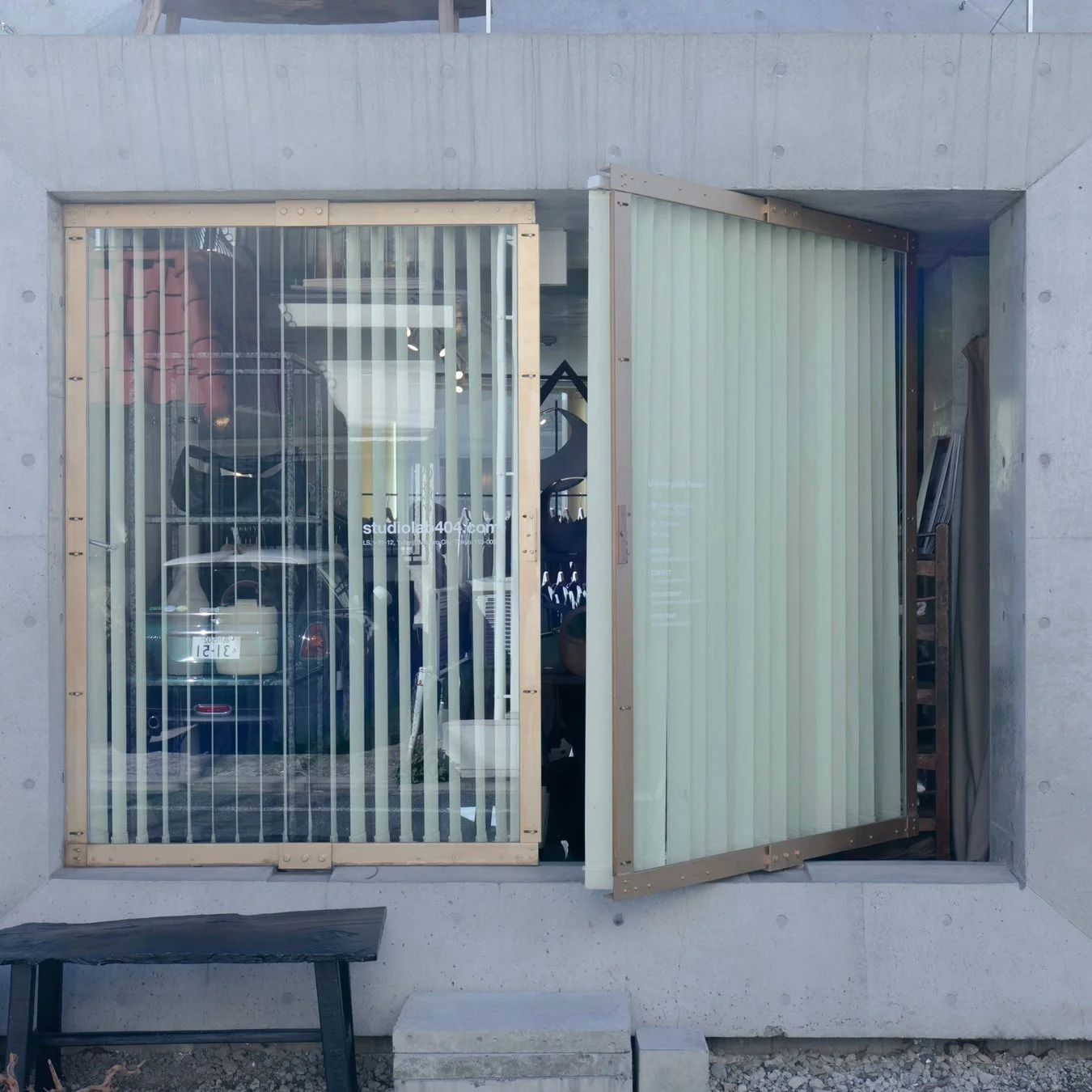 コンクリート造のモダンな建物の外観。縦型ブラインドのある大きな窓の一部が開き、内部には車や商品が見える。