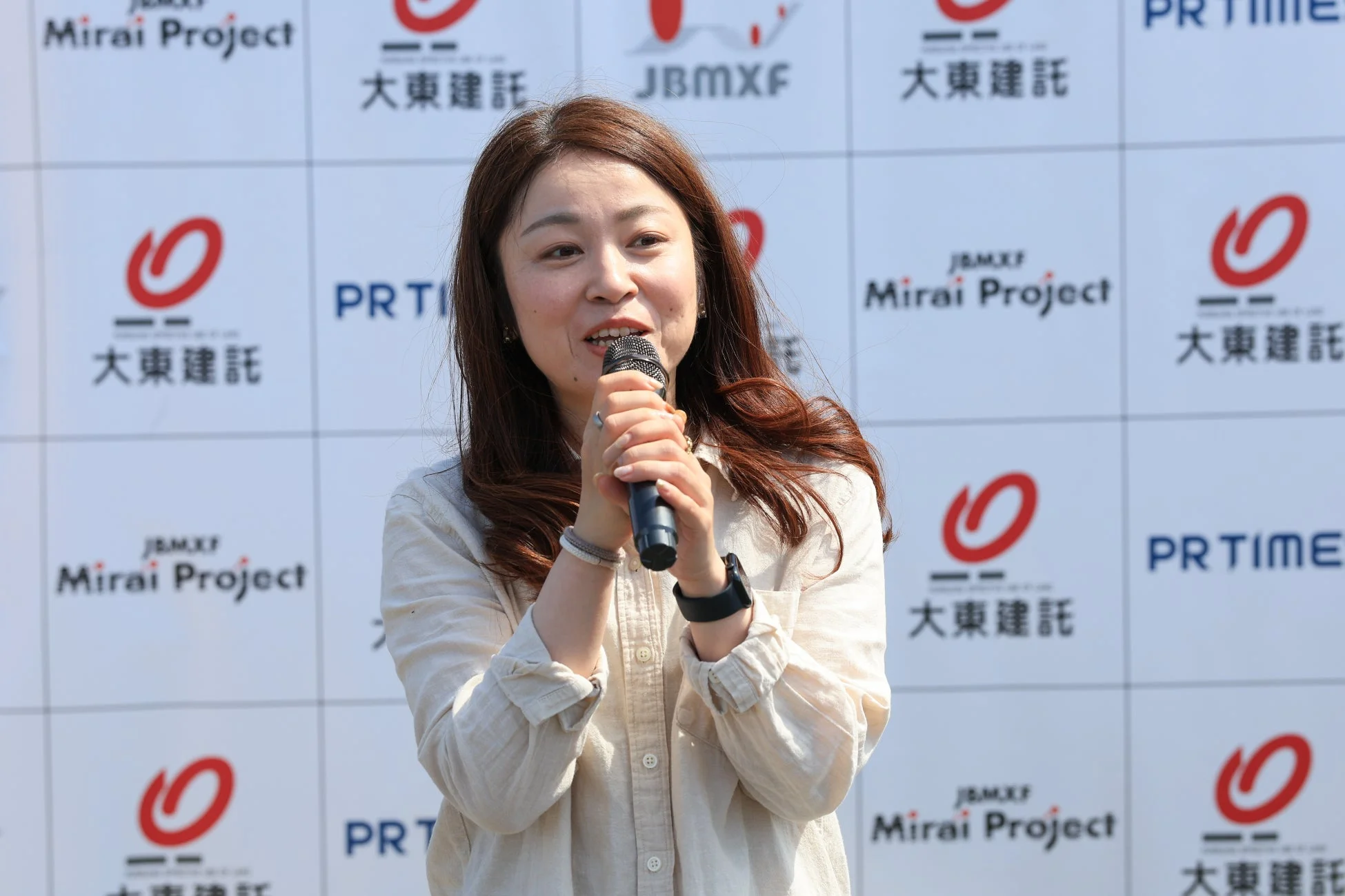 「大東建託」や「Mirai Project」のロゴが並ぶ背景の前で、女性がマイクを手に笑顔でスピーチをしている