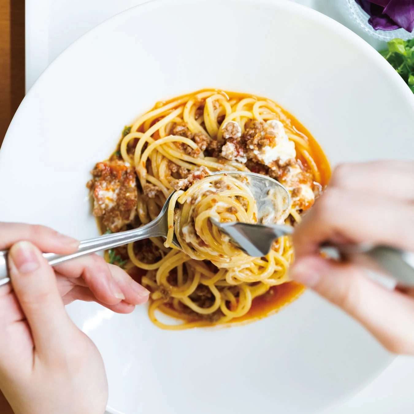 白い皿に盛られたボロネーゼパスタを、スプーンとフォークで巻き取っている様子を上から捉えた写真です。