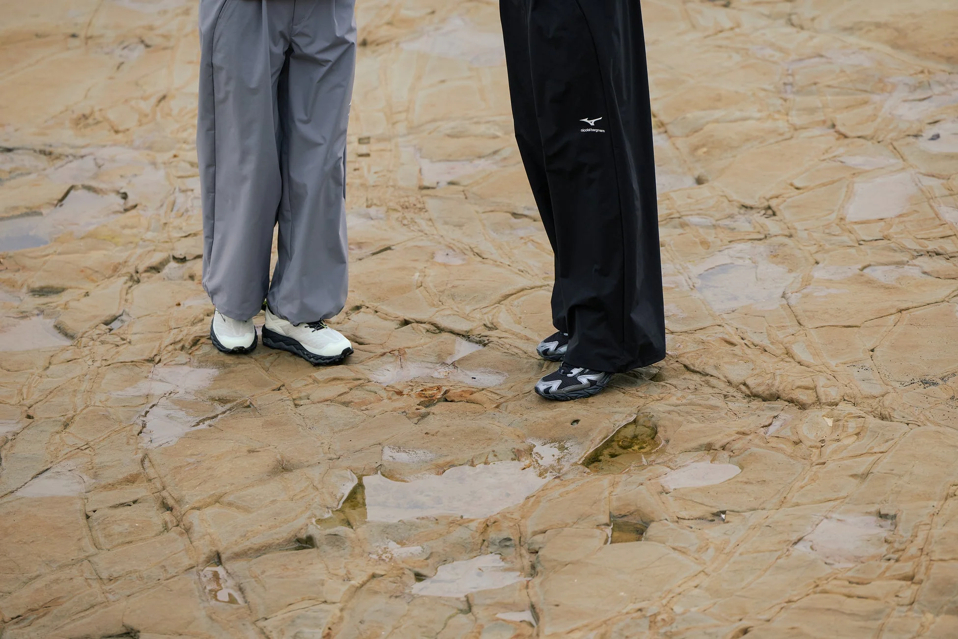 濡れた岩場に立つ2人の足元を写した写真。グレーと黒のパンツにそれぞれ異なるデザインのスニーカーを合わせており、アウトドアカジュアルなスタイリングが特徴です。黒いパンツには「Mizuno」と「nicolai bergmann」のロゴが見えます。